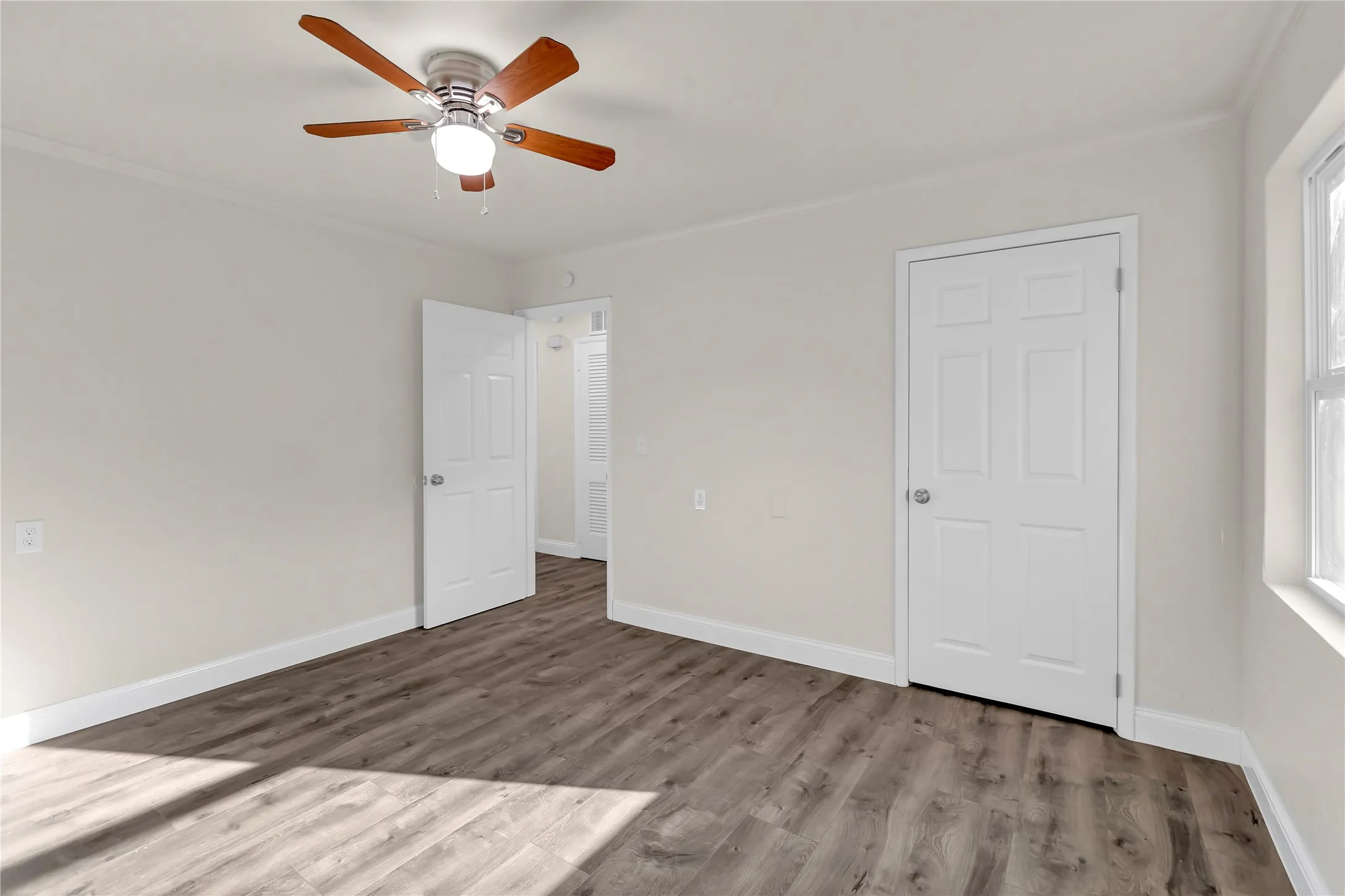 Room featuring light-toned walls, wood-finish flooring, white baseboards, a ceiling fan with light fixture, and two paneled doors