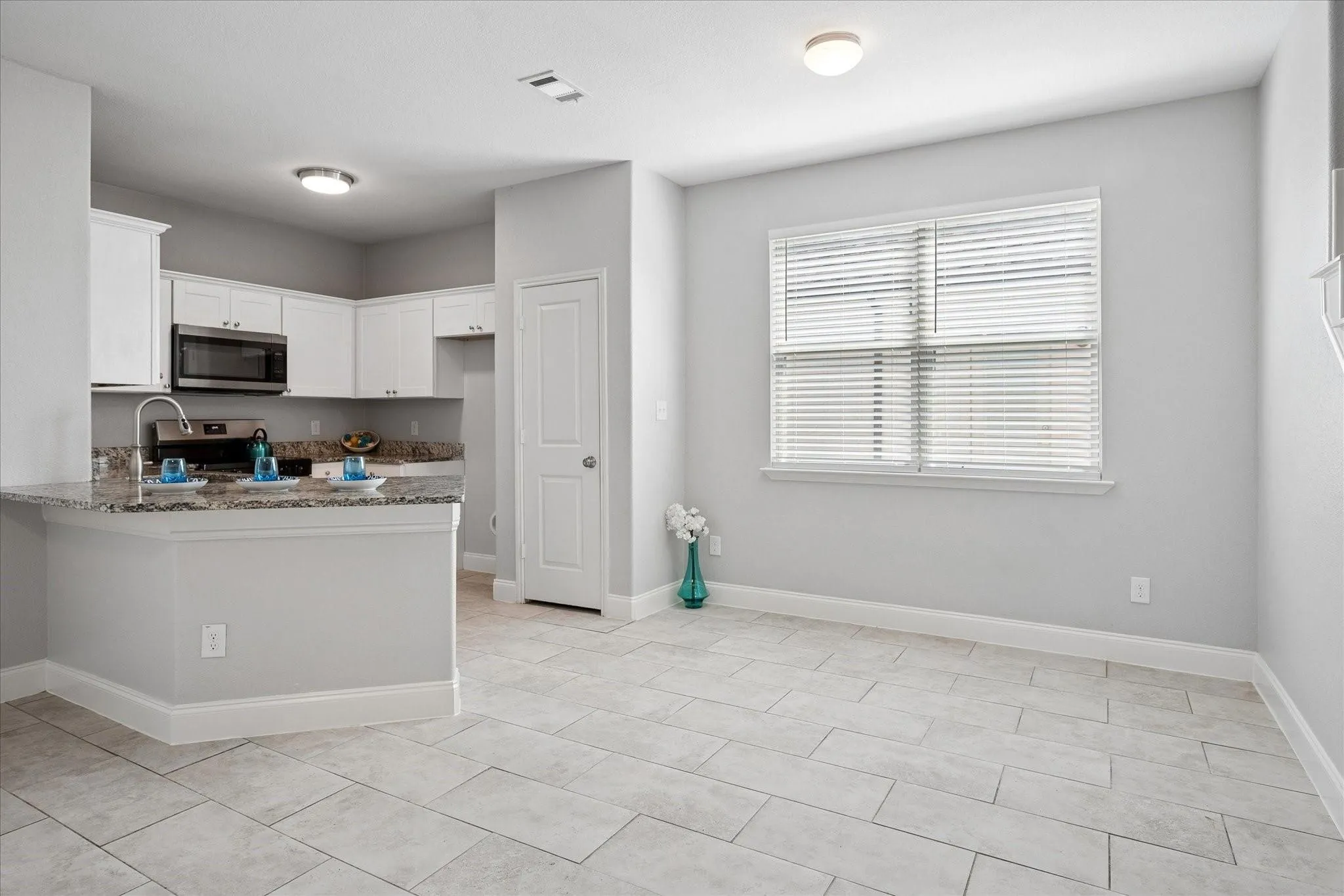 Open-concept living space with large format tile flooring and a kitchen featuring white cabinetry, stainless steel appliances, a granite-finish countertop, and a breakfast bar