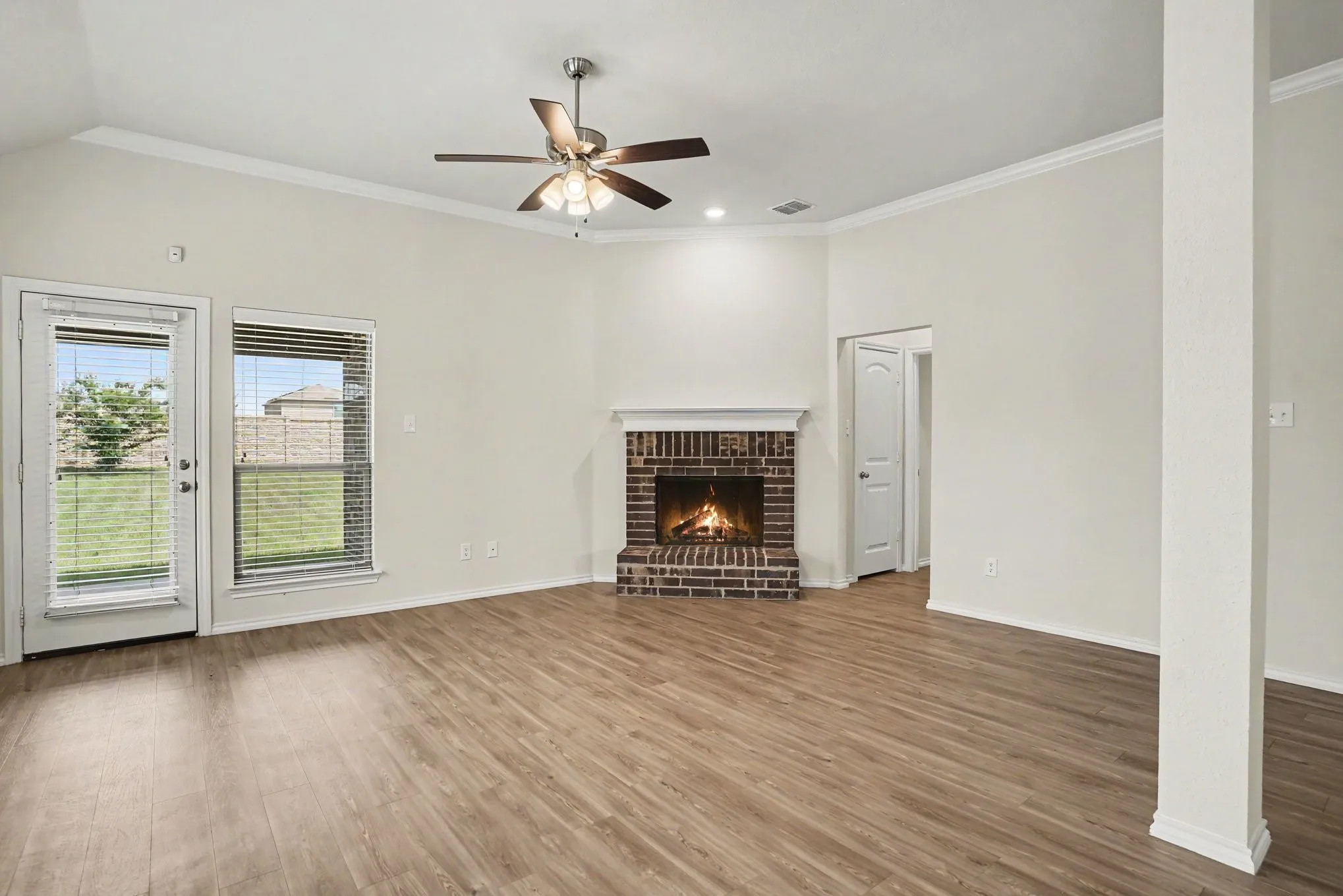 Open living space featuring wood-finish flooring, a brick fireplace with mantel, a ceiling fan, crown molding, and a glass-paneled door opening to a private yard