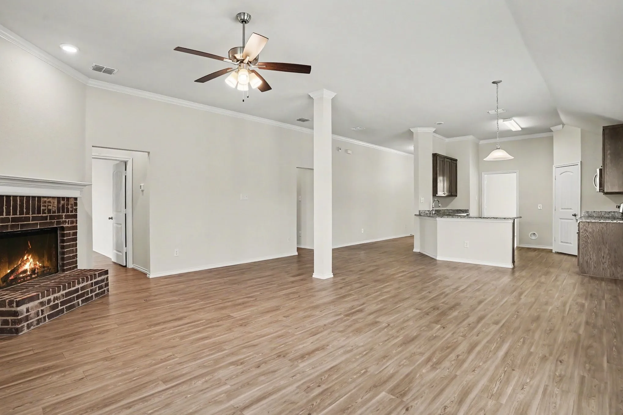 Spacious interior featuring wood-finish flooring, a brick fireplace with a white mantel, crown molding, and a kitchen area with dark cabinetry and granite-style countertops
