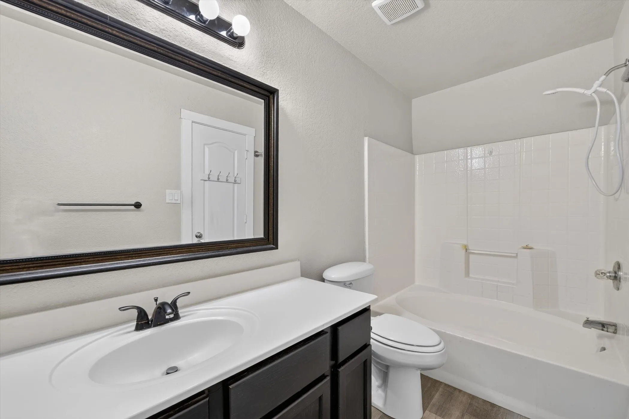 Bathroom featuring a vanity with a white countertop and dark cabinetry, a recessed sink with a matte black faucet, a large framed mirror, a three-light fixture, and a white tub with a shower