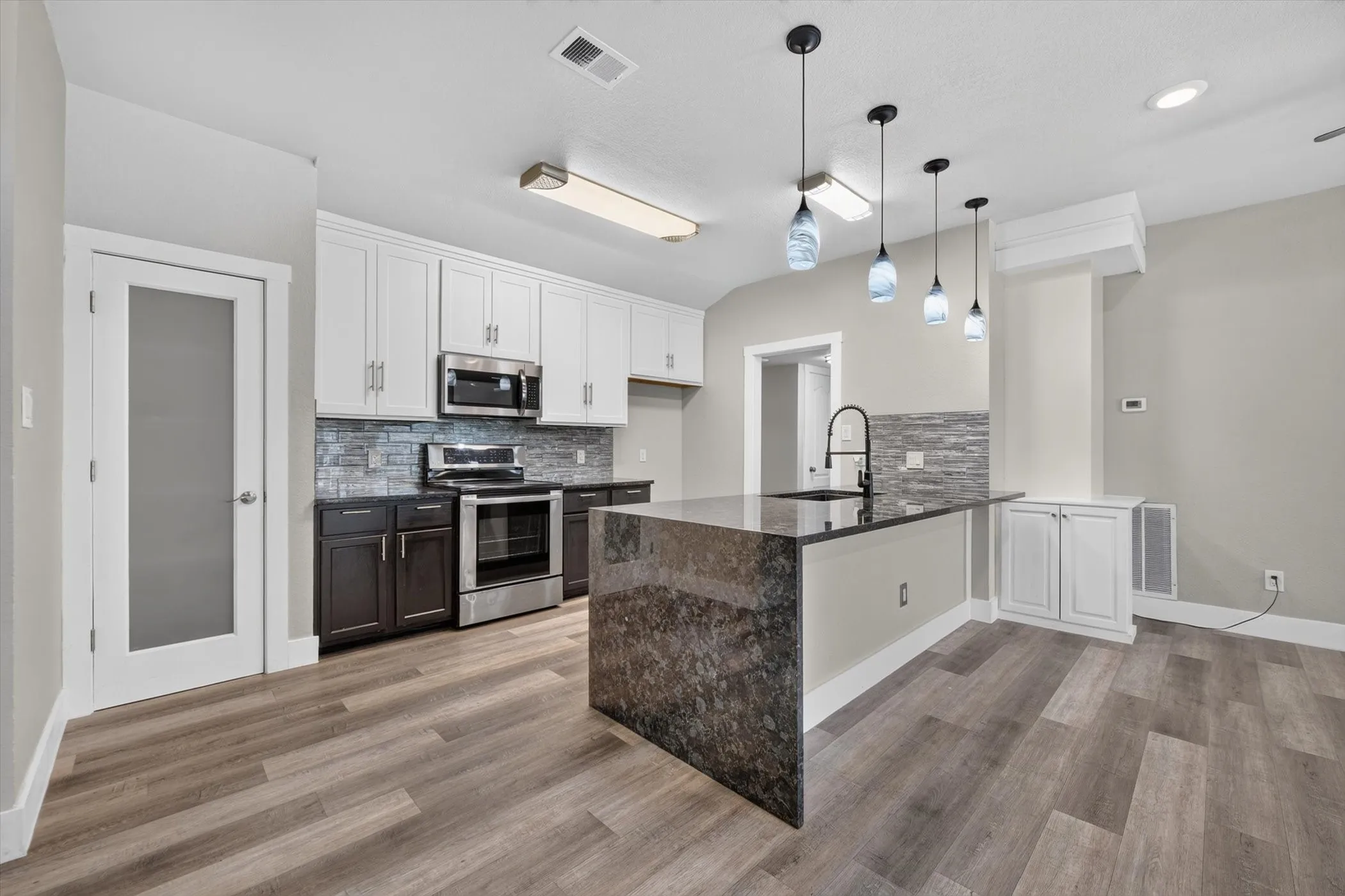 Modern kitchen featuring a waterfall island with a dark stone countertop, pendant lighting, stainless steel appliances, two-tone cabinetry, and wood-finish flooring