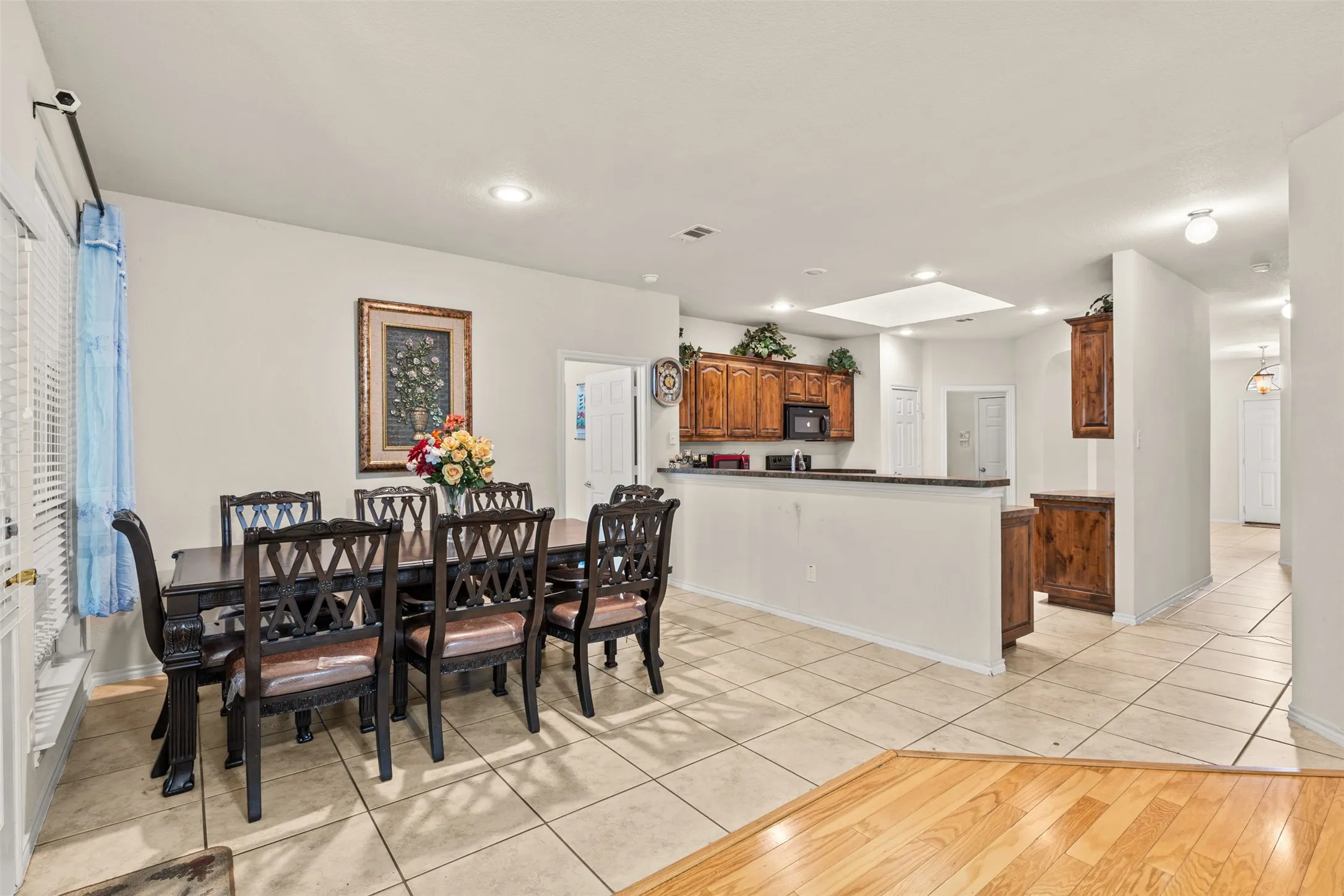 Open-concept interior featuring light-colored tile flooring, warm wood cabinetry, recessed lighting, and a kitchen pass-through with a dark countertop