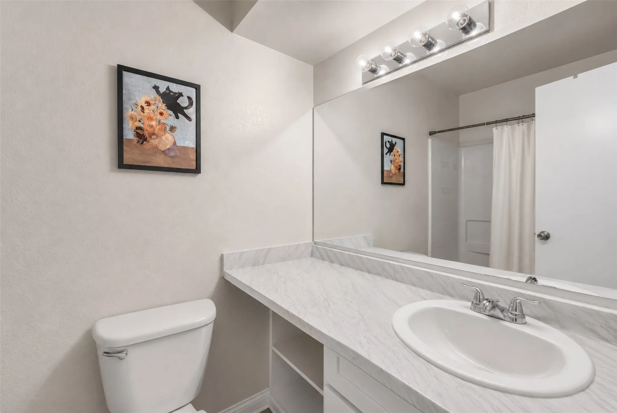 Bathroom featuring a white toilet, an expansive vanity with a white-patterned countertop, an integrated sink with a chrome faucet, and a large mirror with a four-bulb light fixture