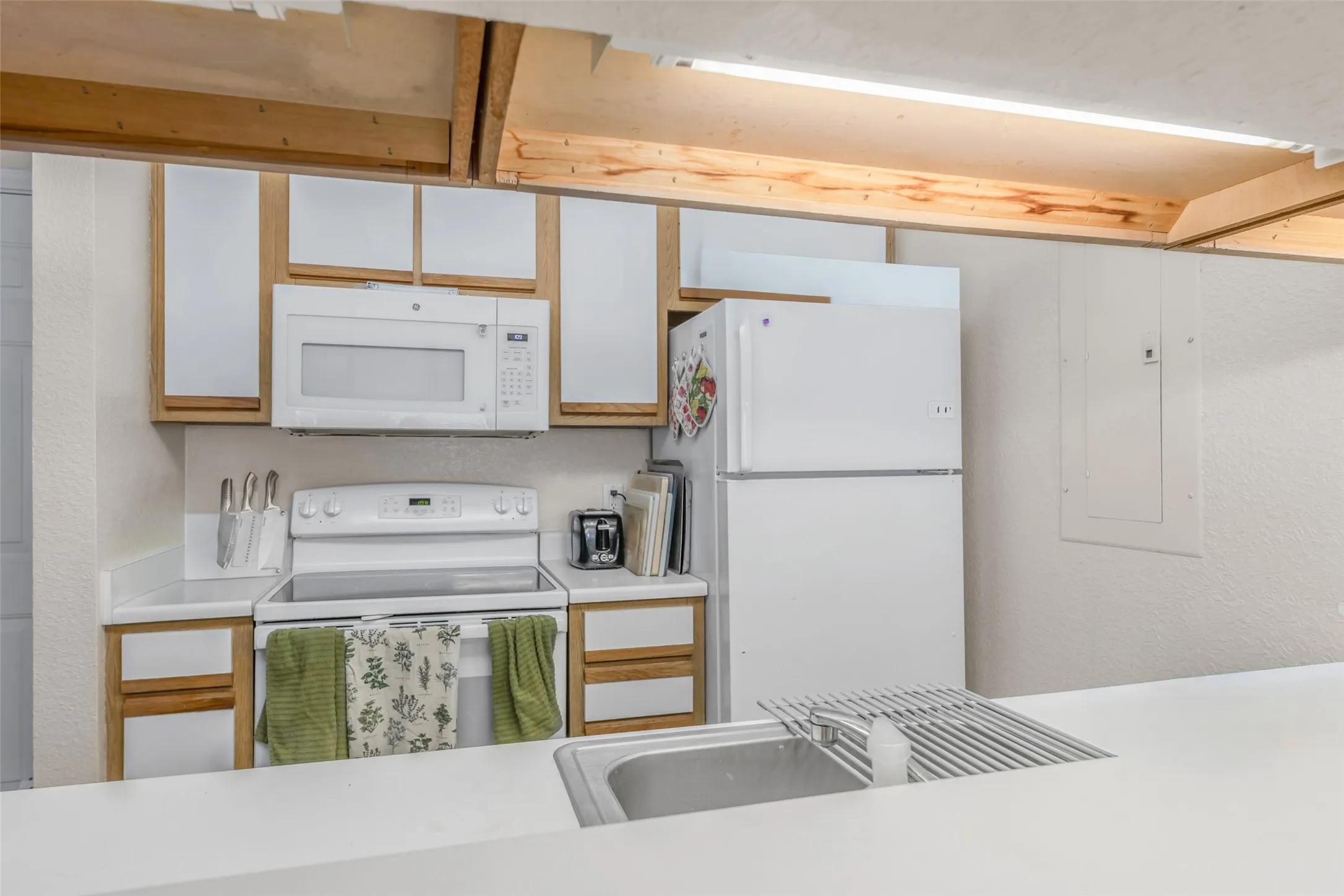View of kitchen from living area featuring a built-in microwave, white electric range, white refrigerator, wood-finish cabinetry with white panels, and a stainless steel sink set into a white countertop