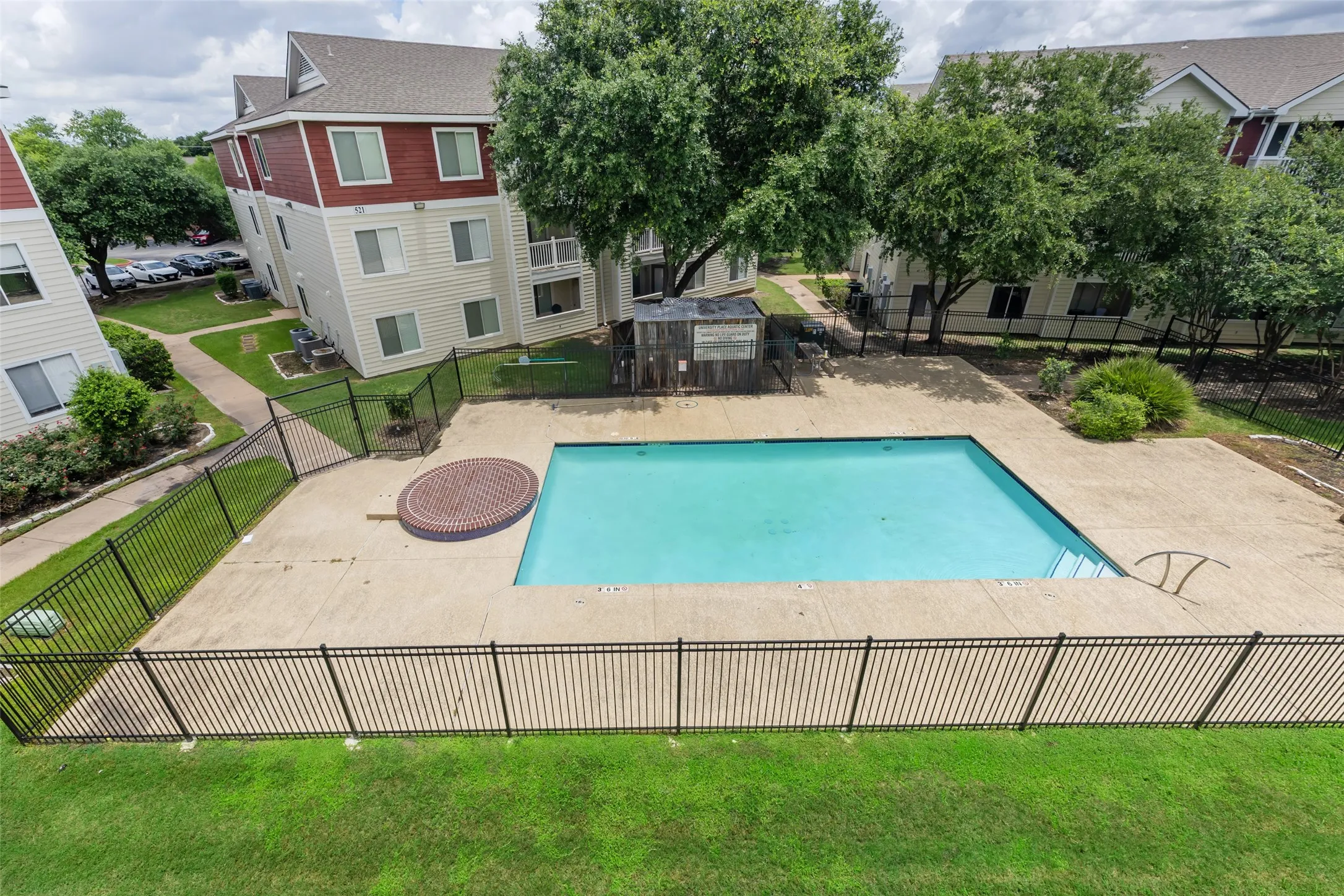 Fenced outdoor swimming pool with surrounding concrete decking, complemented by a circular brick-patterned hardscape feature