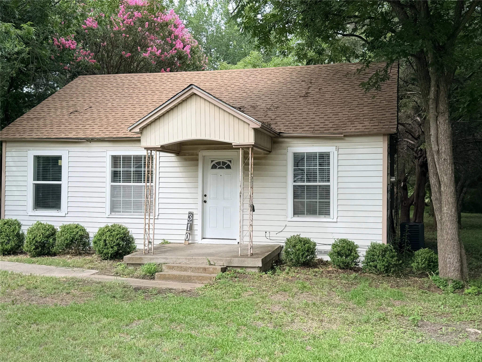 White siding exterior with a prominent front porch, brown shingle roof, and established landscaping