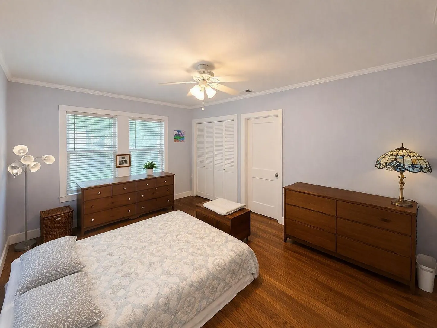 Light-filled room featuring wood-finish flooring, two windows with blinds, a ceiling fan, and white trim molding