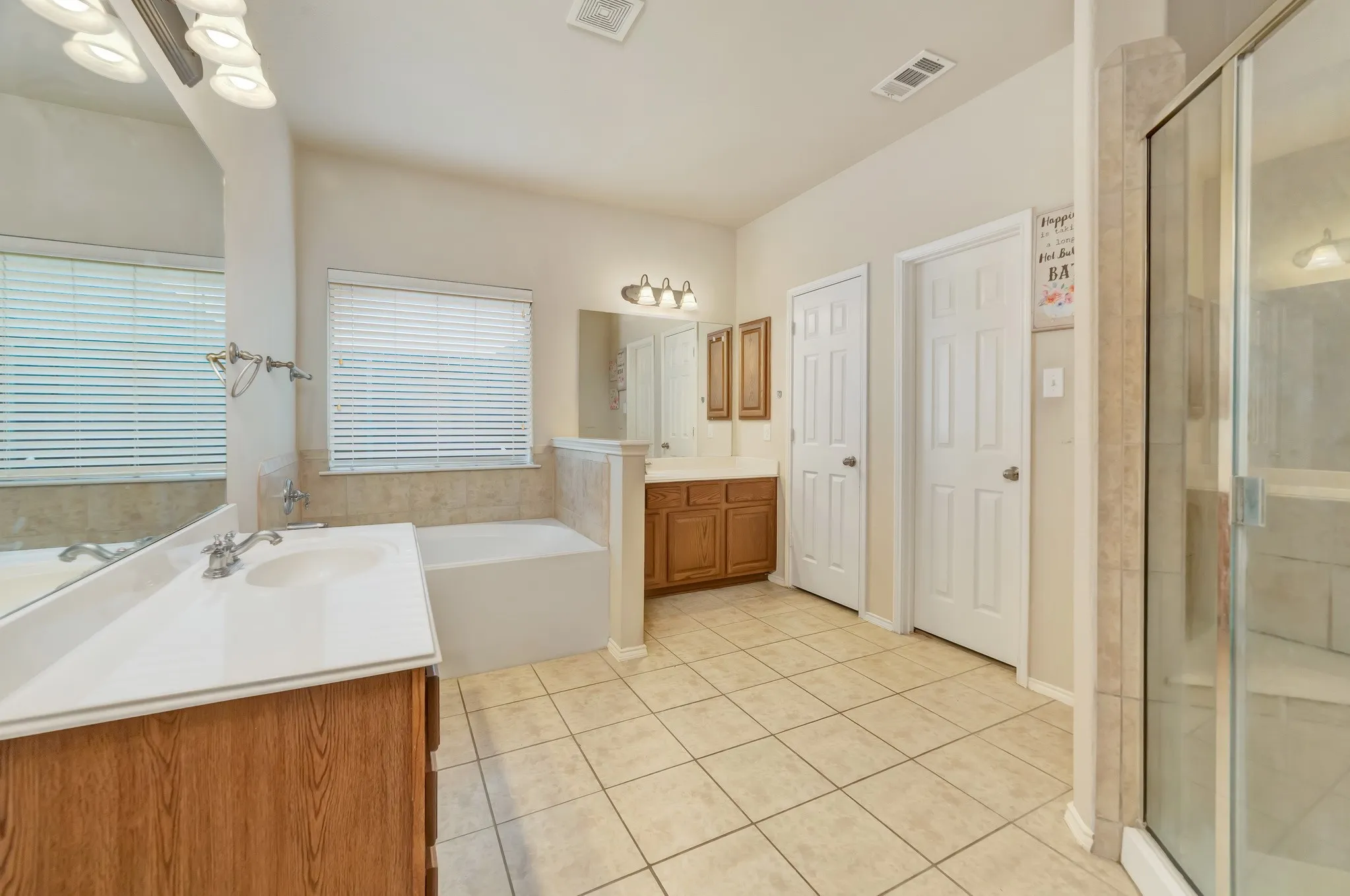 Spacious bathroom featuring a built-in tub with tile surround, a separate glass-enclosed shower, dual wood-finish vanities with white countertops, and light-toned tile flooring
