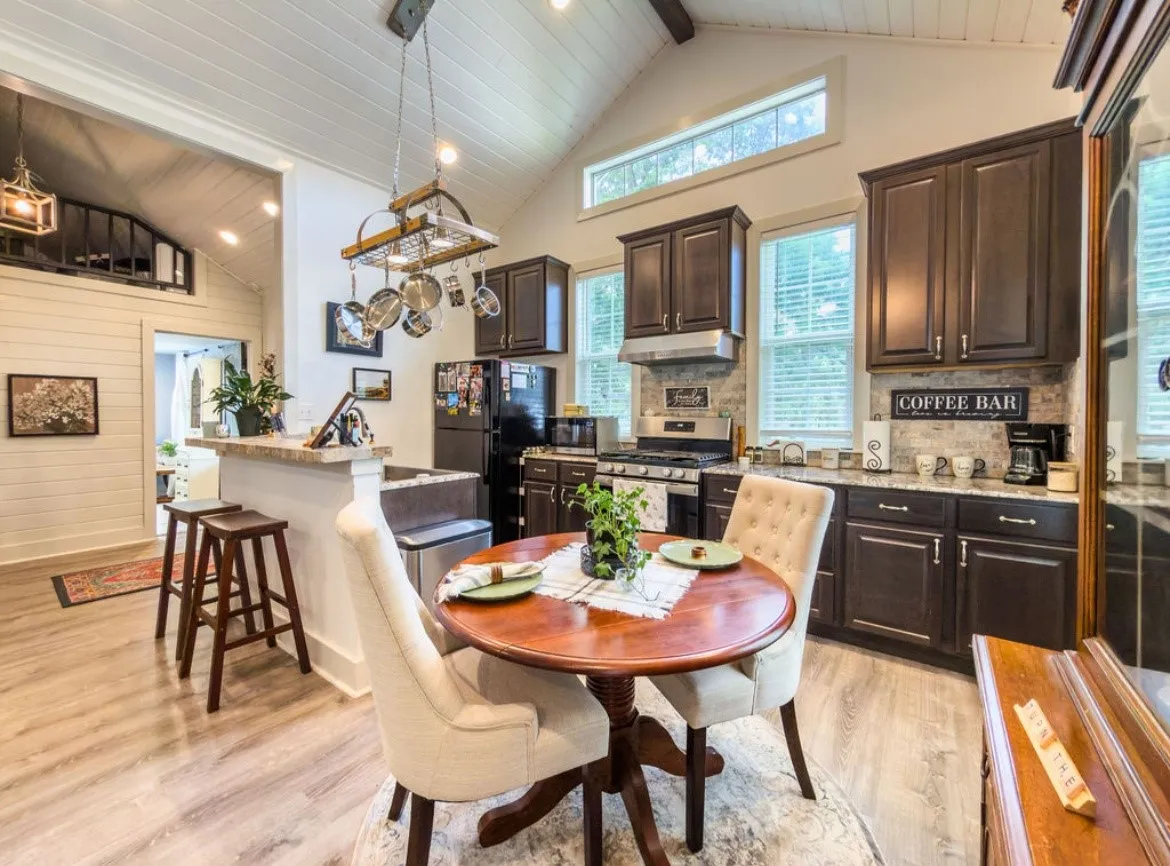 Open-concept kitchen and dining area featuring vaulted ceilings with exposed beams, wood-finish flooring, dark wood cabinetry, a subway tile backsplash, and a breakfast bar