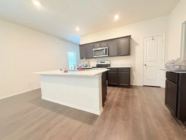 Kitchen featuring dark wood cabinetry, stainless steel appliances, a white island with an integrated sink, light countertops, and wood-finish flooring