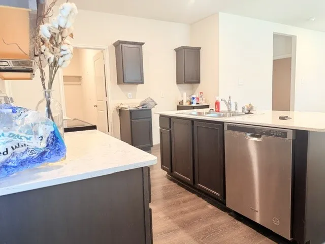Kitchen area featuring dark wood-finish cabinetry, light-toned countertops, a stainless steel dishwasher, a stainless steel sink with a chrome faucet, and wood-finish flooring