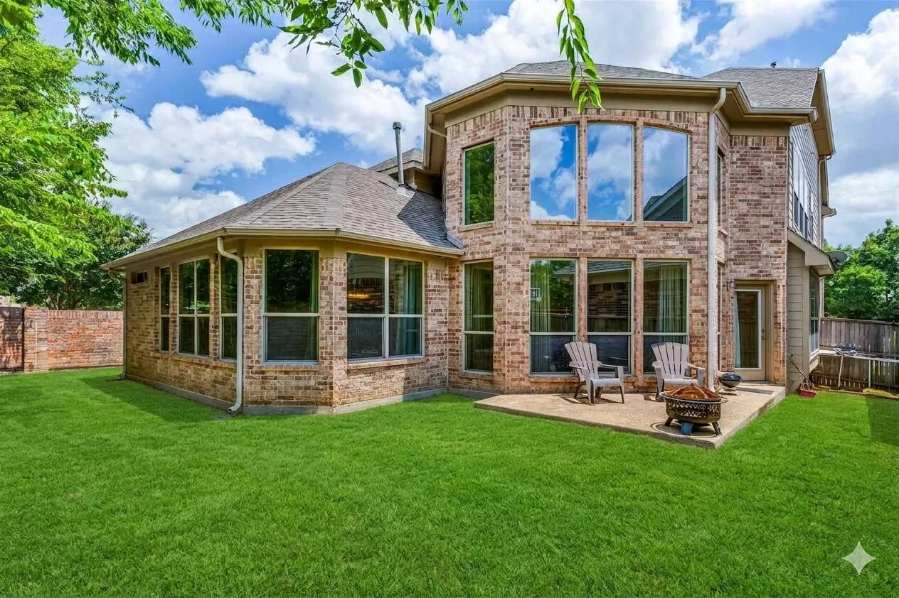 Brick exterior with a multi-level roofline, featuring numerous windows, an expansive green lawn, a stone patio, and a privacy fence