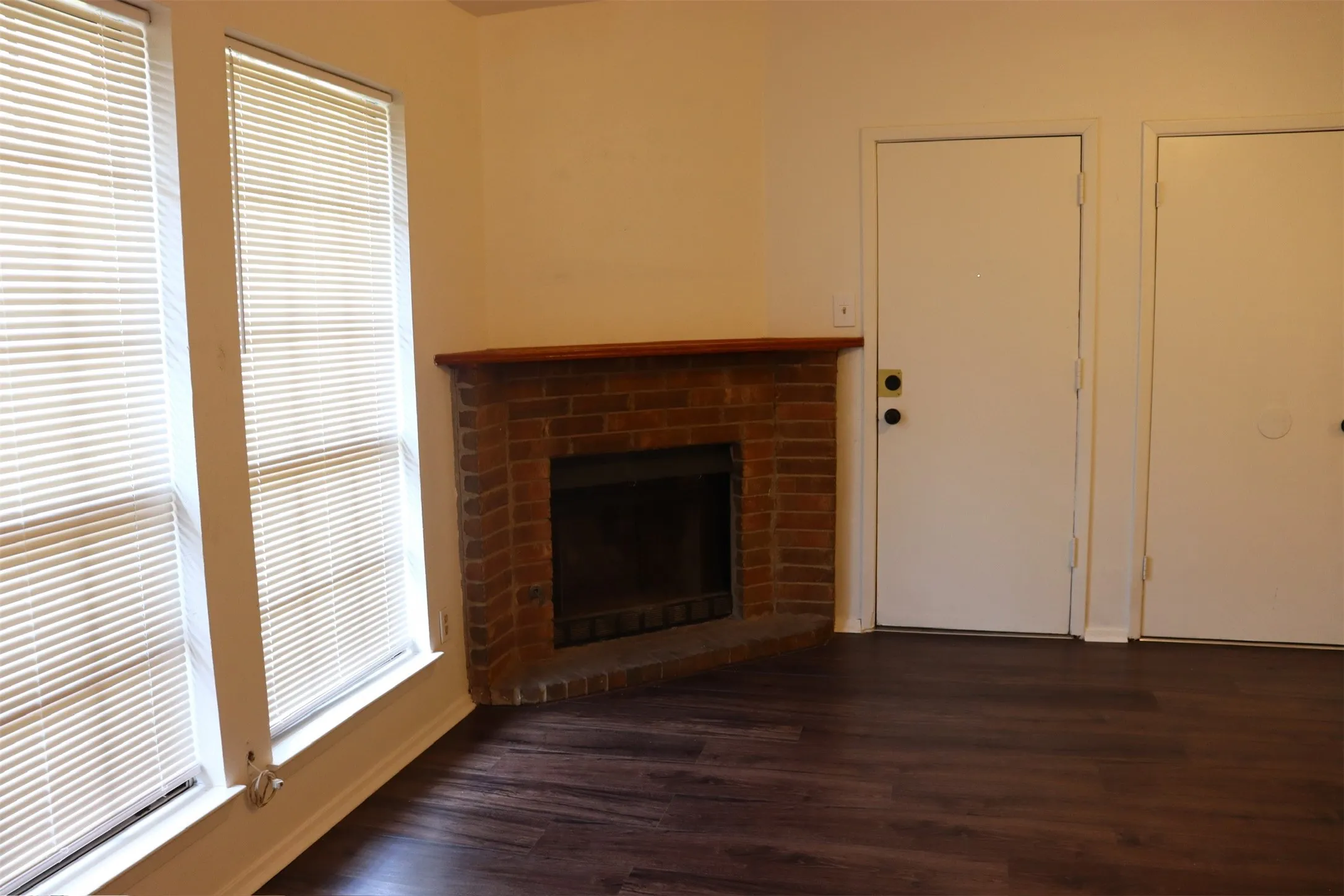 Living area featuring a corner brick fireplace with a wood mantel, dark wood-finish flooring, and two windows with blinds