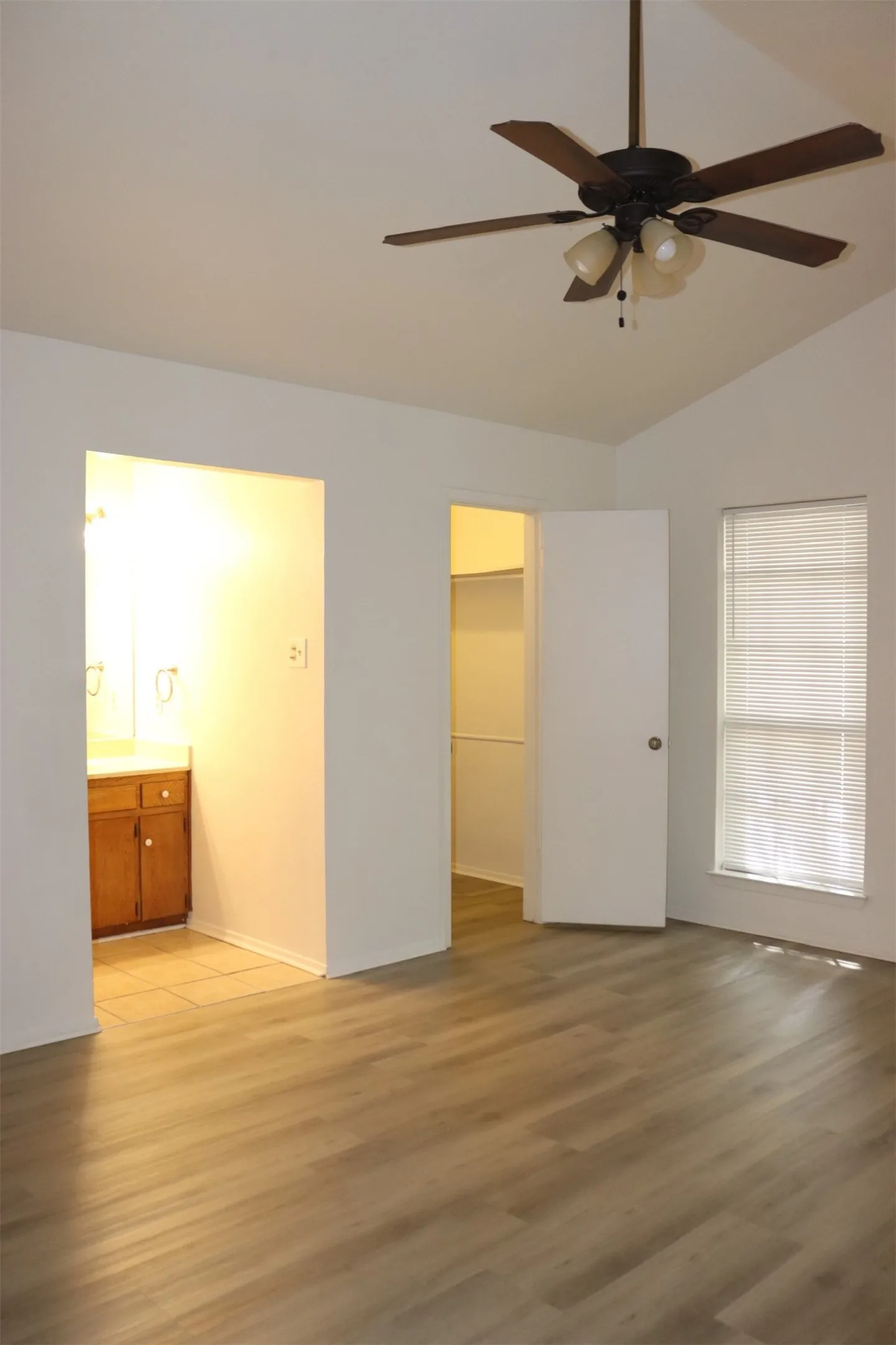 Spacious room featuring vaulted ceilings, wood-finish flooring, a ceiling fan with light kit, a window with blinds, and an entryway to a bathroom with a wood vanity
