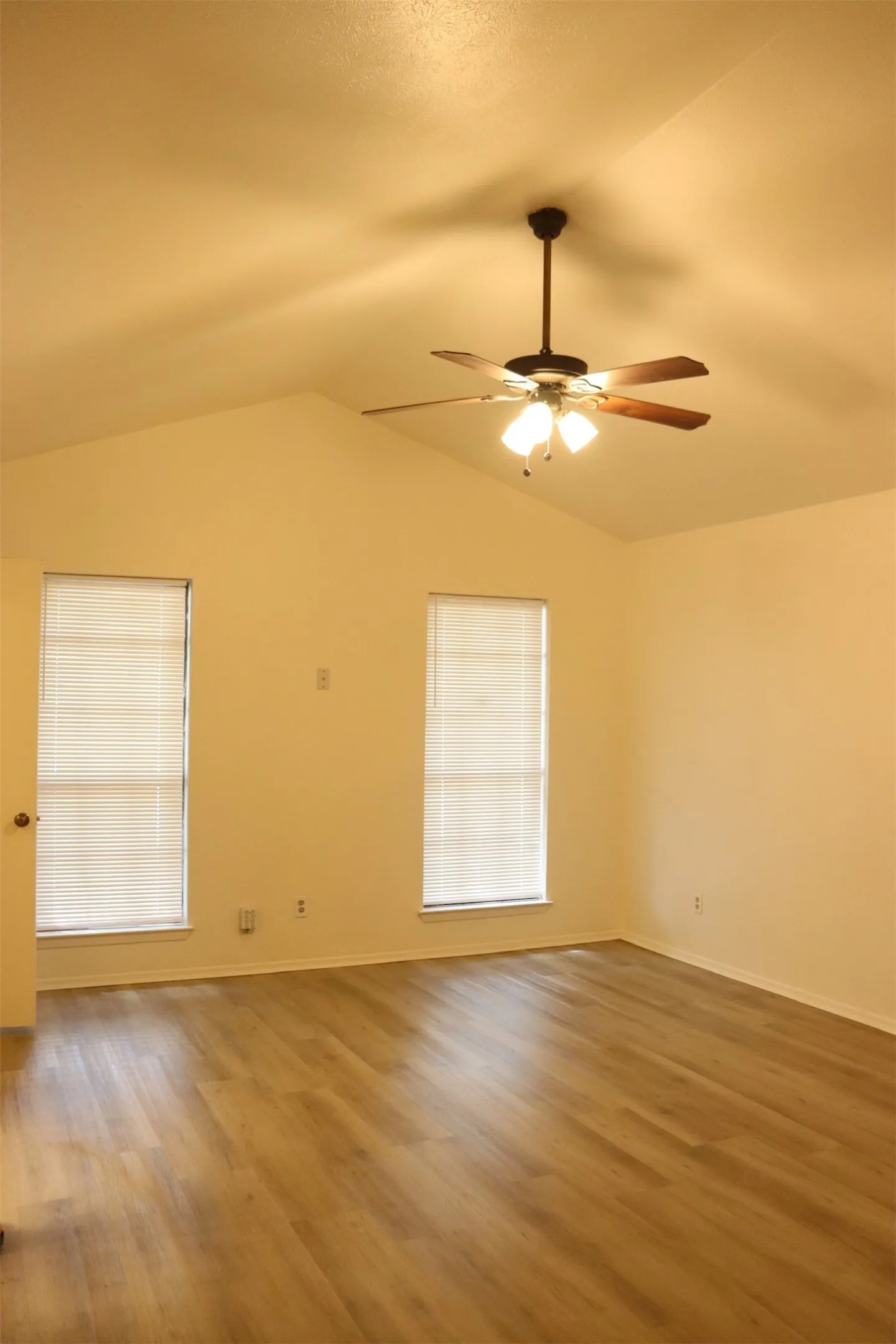 Spacious room featuring vaulted ceilings, wood-finish flooring, two large windows with blinds, and a ceiling fan with integrated lighting