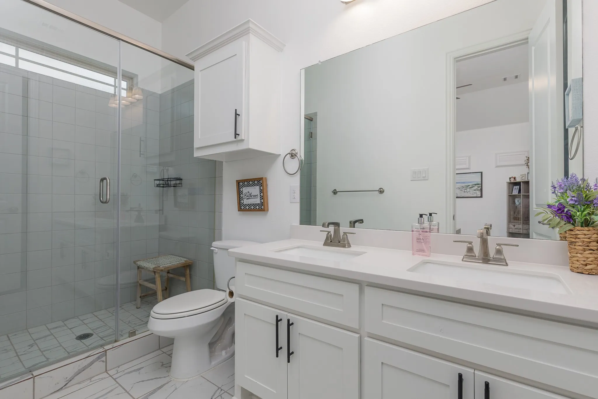 Bathroom featuring a dual vanity with undermount sinks and brushed nickel faucets, a large wall-mounted mirror, white shaker-style cabinetry with black hardware, a glass-enclosed shower, and white tile flooring