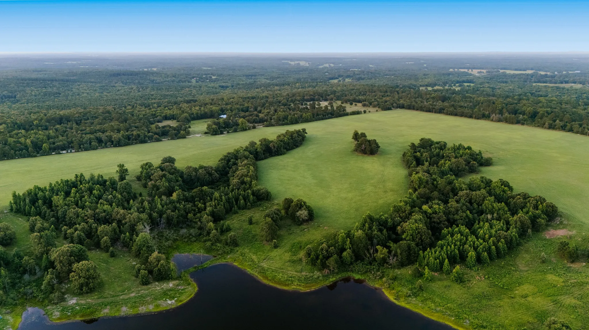 Expansive verdant landscape featuring a substantial body of water, extensive wooded areas, and open fields under a clear sky