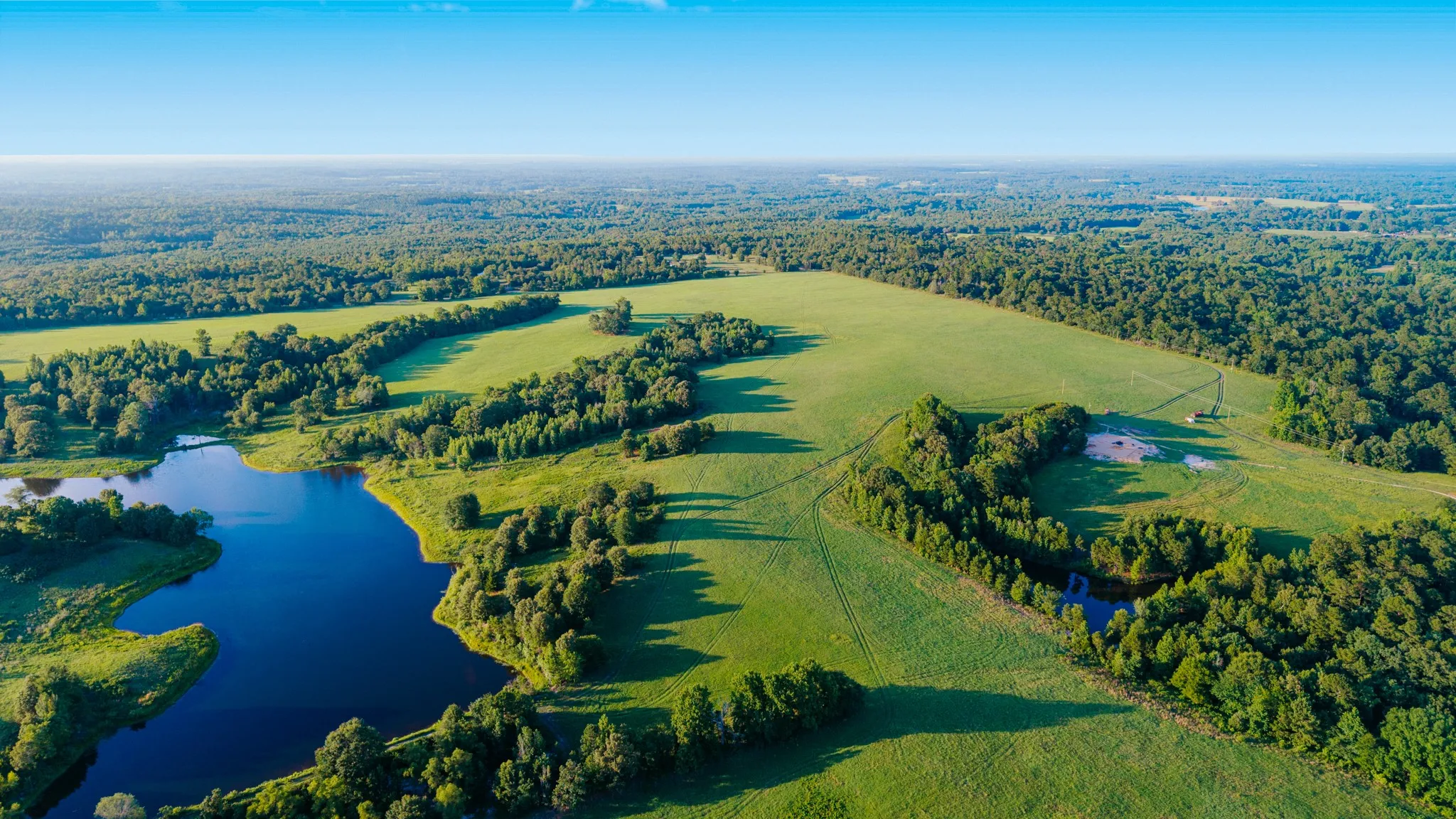 Expansive land featuring a large pond, open fields, and mature tree lines