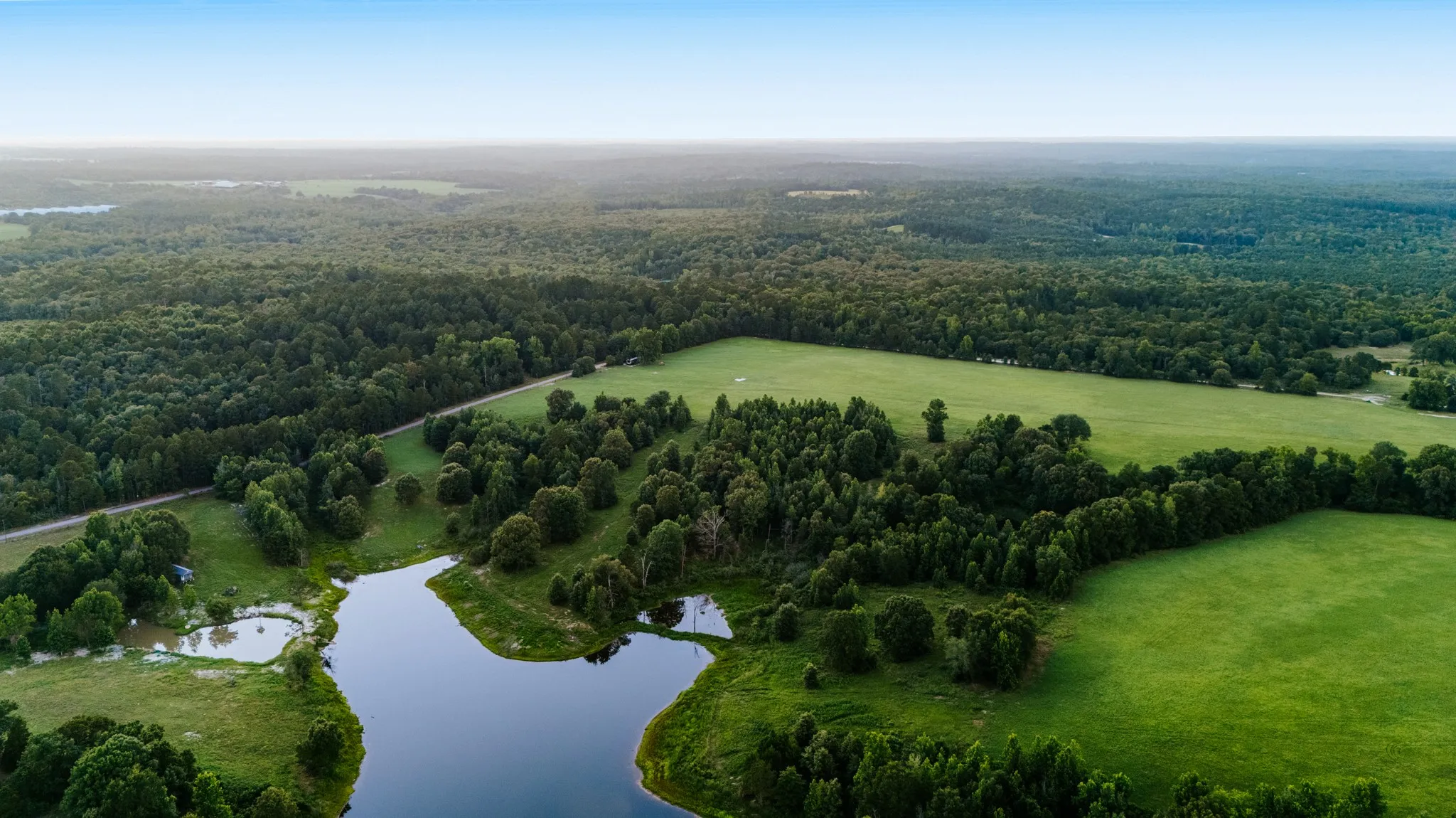 Expansive property featuring a large body of water, lush green fields, mature trees, and dense woodland