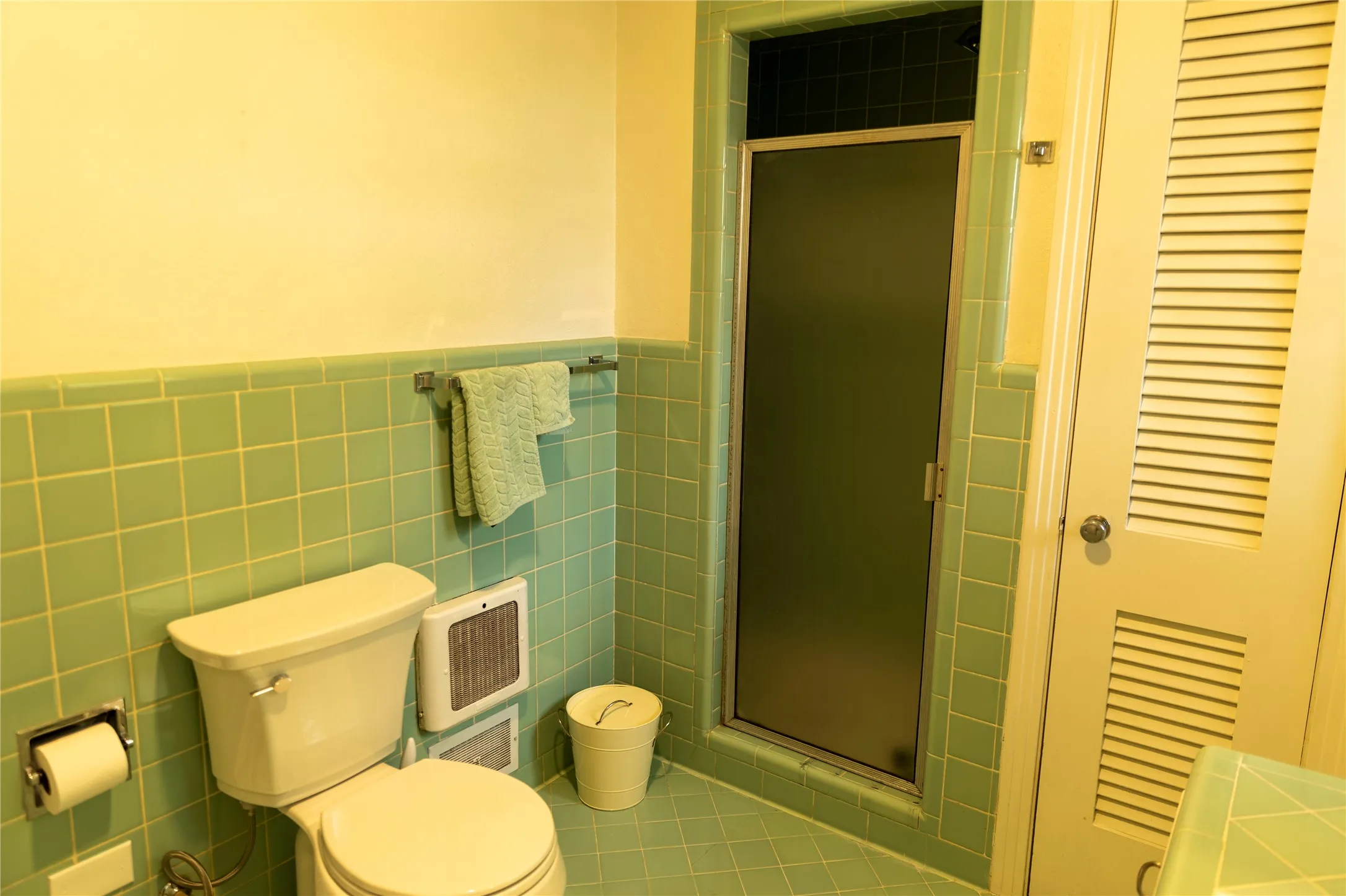 Bathroom with square green wall and floor tiling, a white toilet, and a stand-up shower with a frosted glass door
