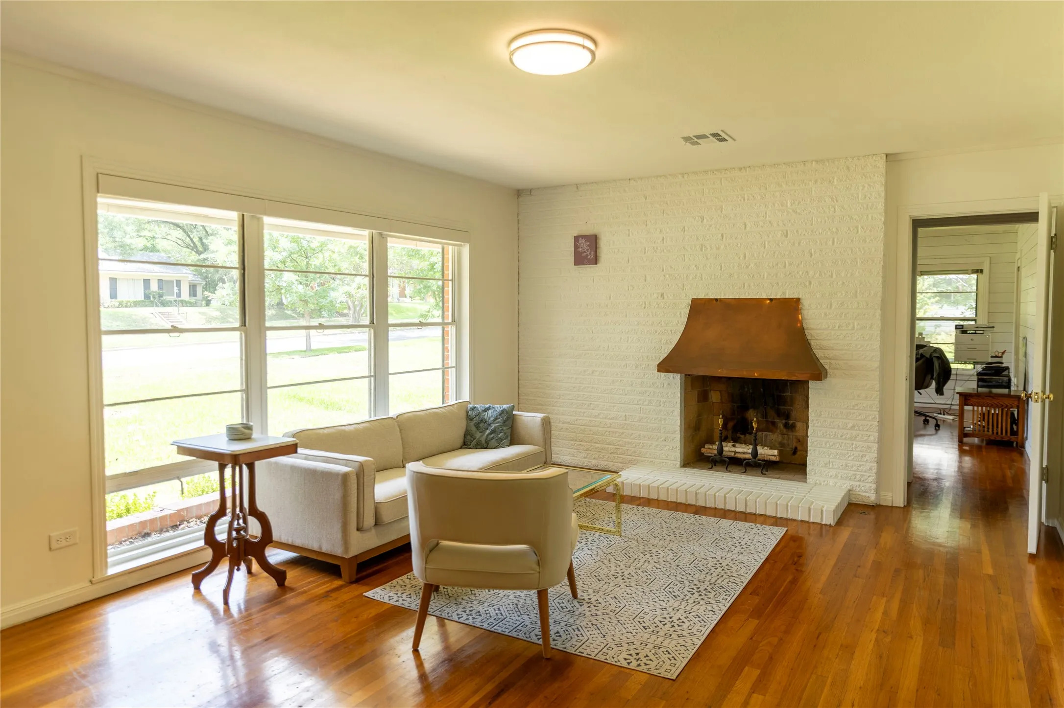 Living area featuring hardwood flooring, a white-painted brick fireplace with a copper hood, and a large window overlooking exterior landscaping