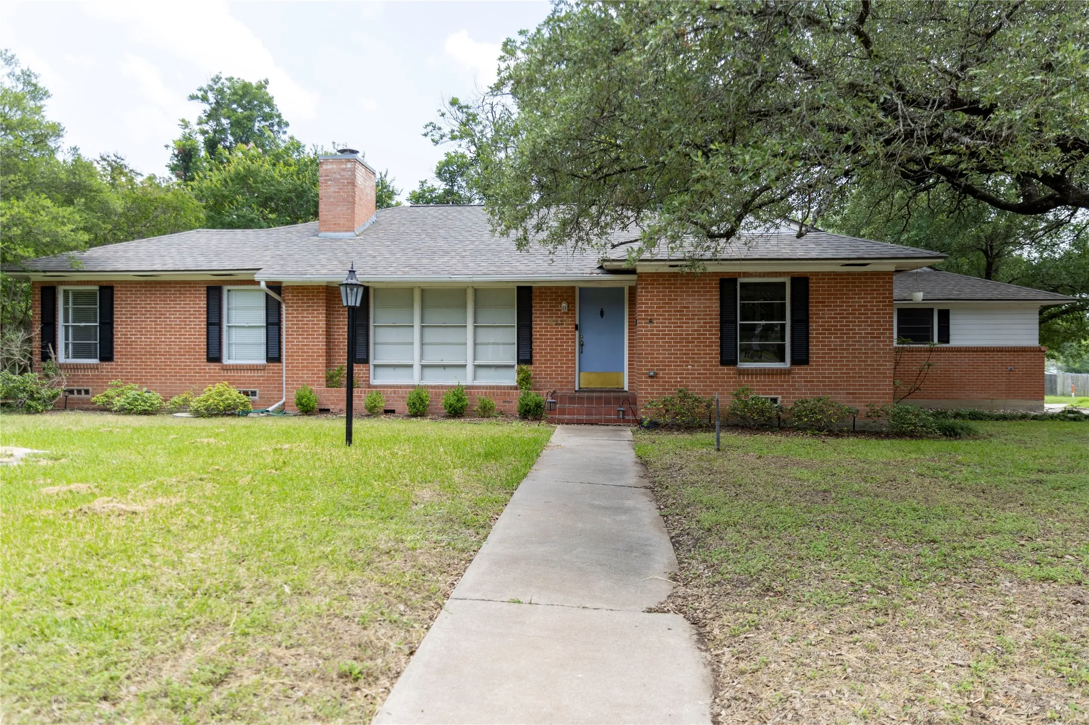Brick exterior featuring a light blue entry door, black window shutters, a front walkway, a covered porch, and a mature tree