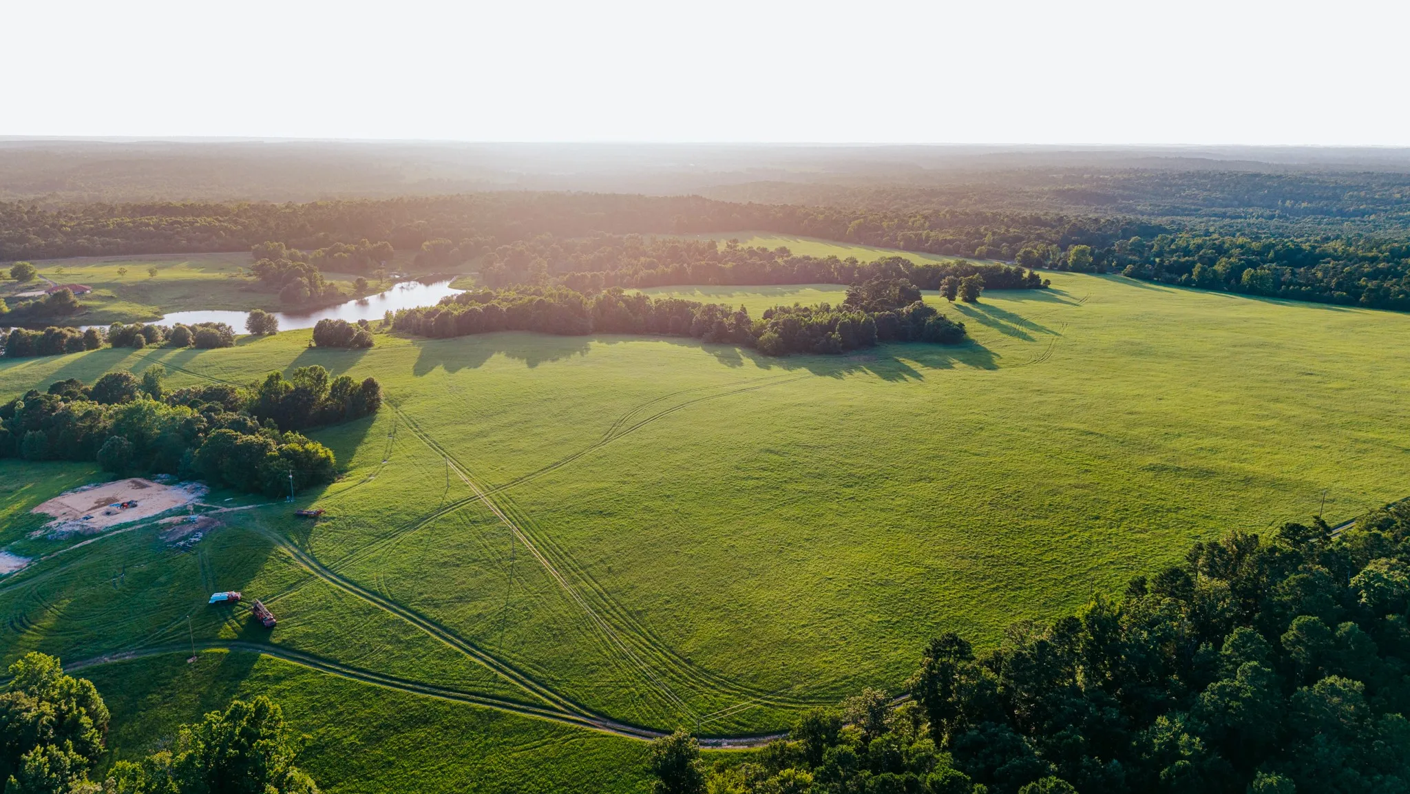 Expansive green acreage featuring a tranquil pond, dense surrounding woodlands, and clear skies