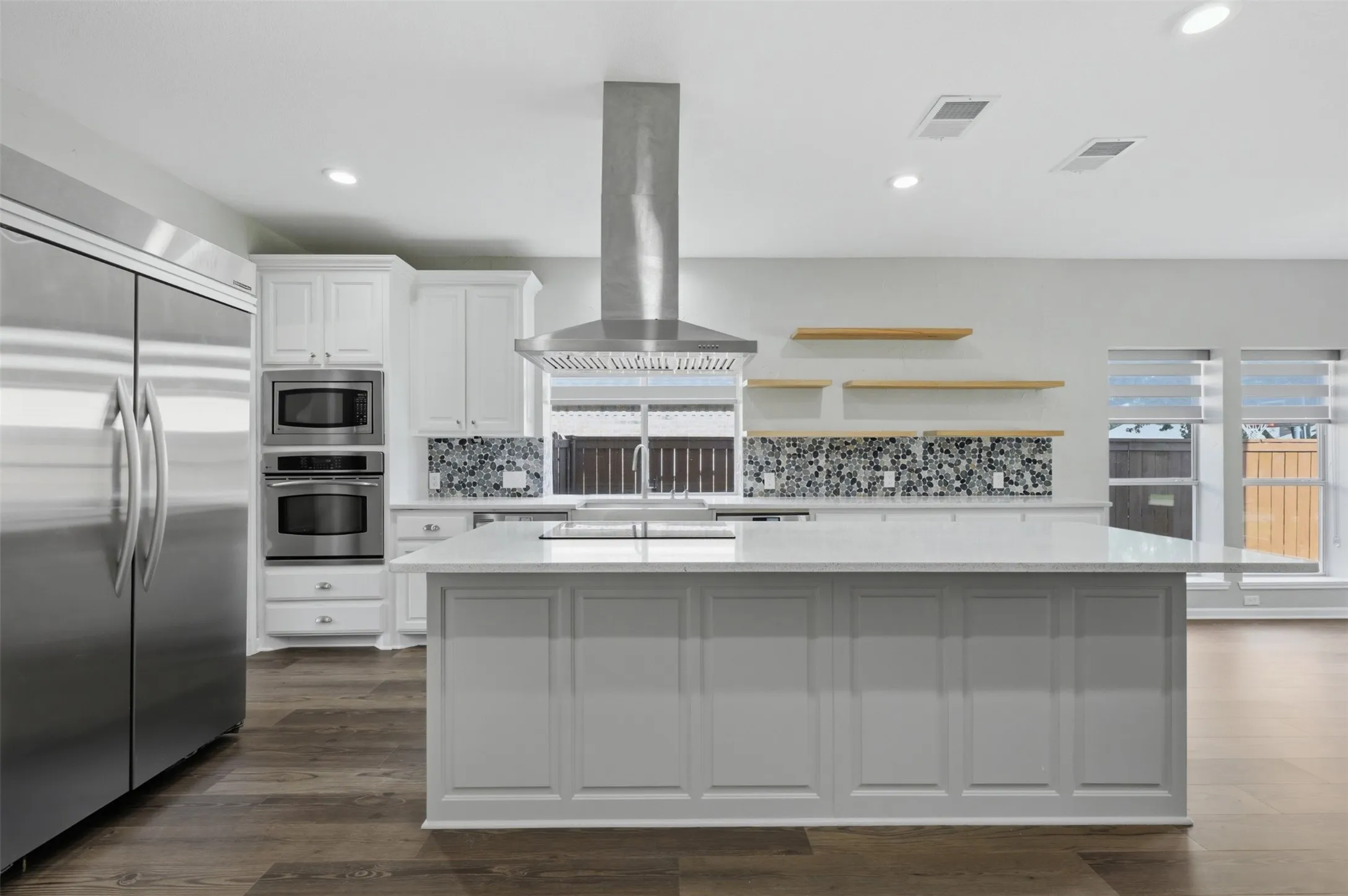 Spacious kitchen featuring a central island with white paneling, stone countertops, stainless steel appliances, a mosaic tile backsplash, and wood-finish flooring