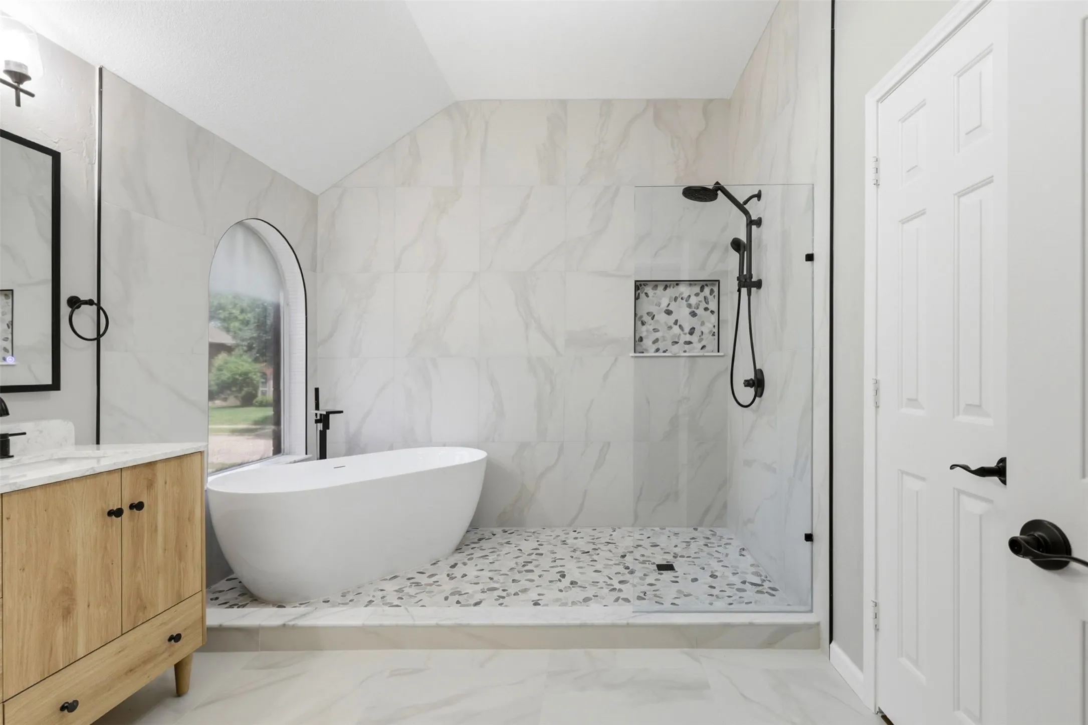 Modern bathroom featuring a freestanding soaking tub, arched window, walk-in shower with stone tile floor, natural wood-finish vanity with stone countertop, and matte black fixtures
