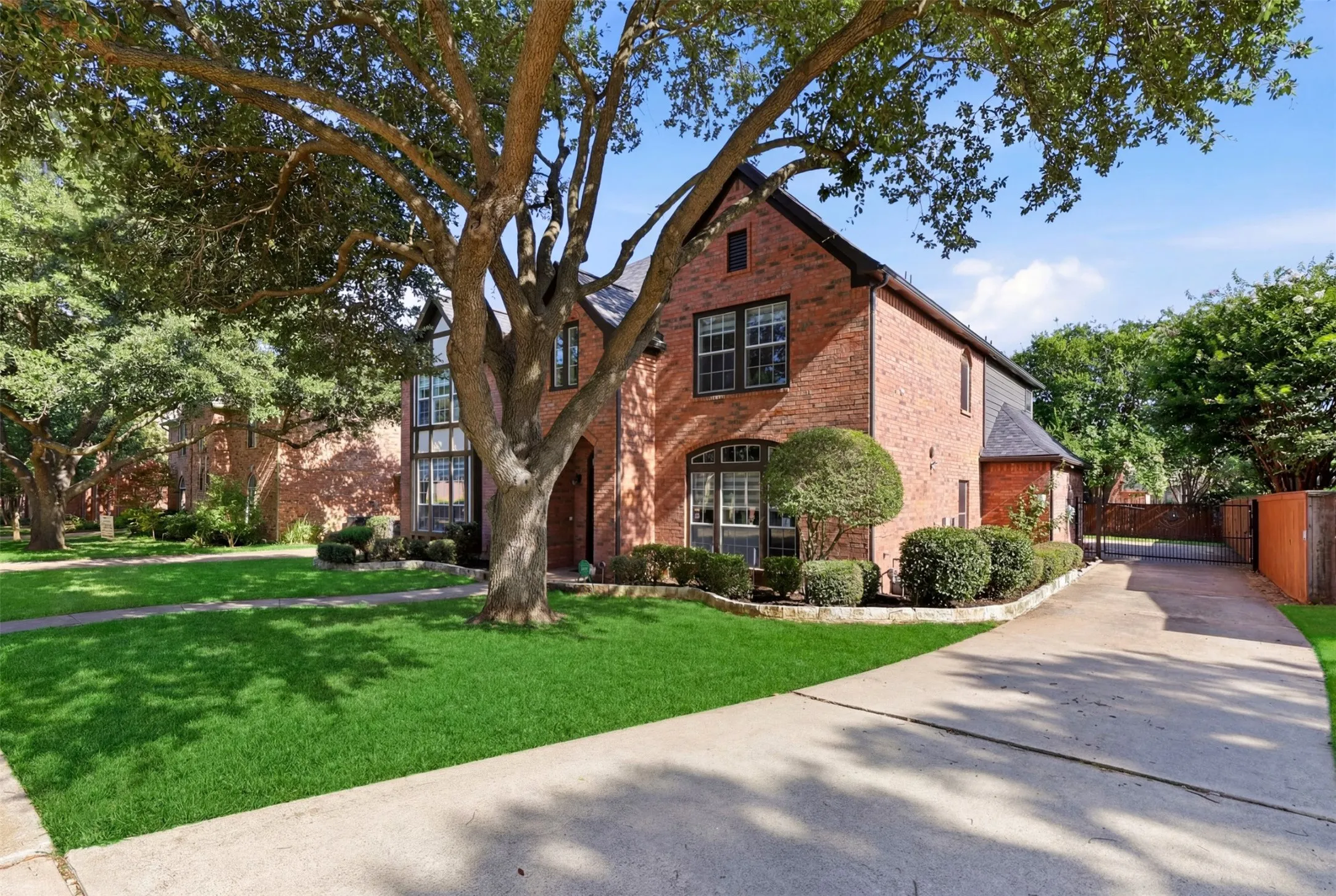 Brick residence featuring a gabled roofline, established trees, a manicured lawn, a concrete driveway, and mature landscaping