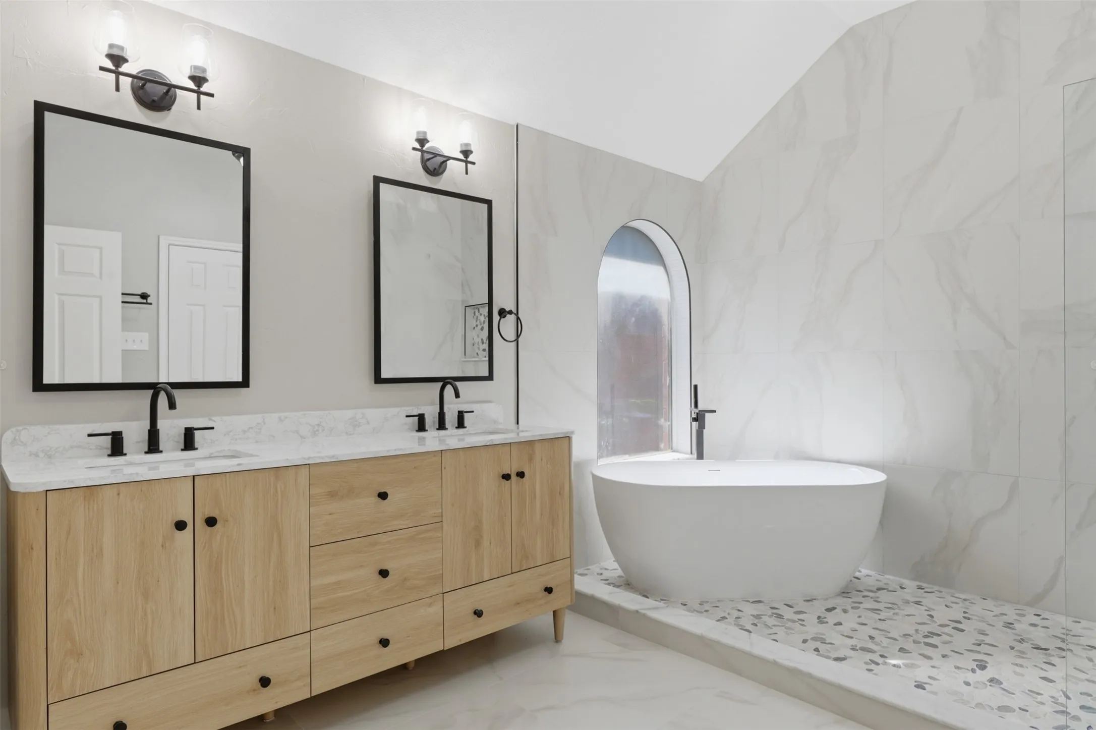 Freestanding soaking tub with arched window, complemented by a dual vanity featuring light wood-finish cabinetry, stone countertop, and matte black fixtures