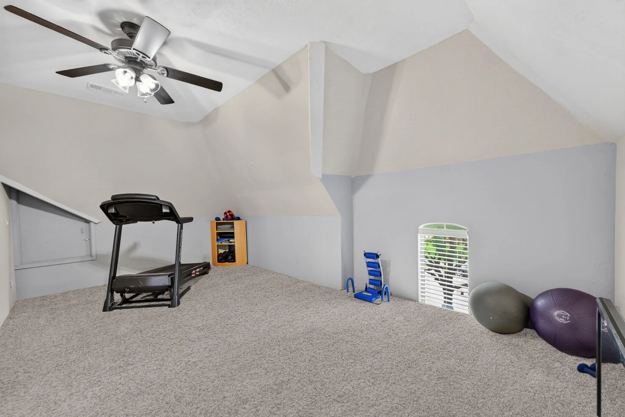 Carpeted room with sloped ceilings, featuring a ceiling fan with integrated lighting, a small arched window with blinds, and light-toned walls