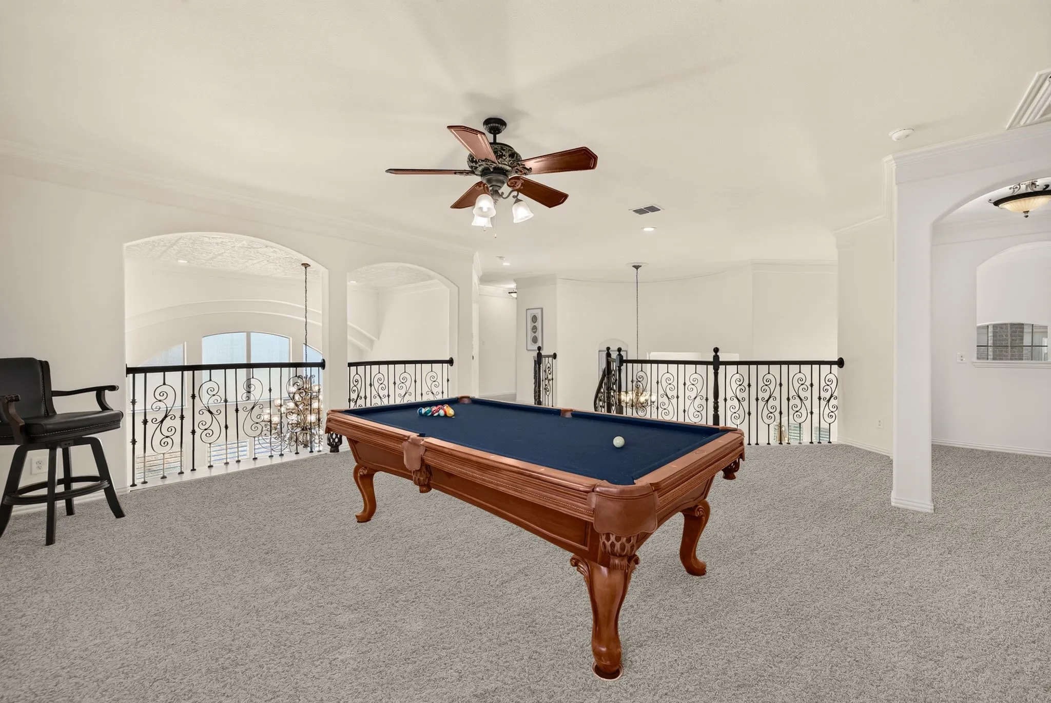Spacious loft area featuring light gray carpeting, decorative wrought iron railings, an arched entryway, and a ceiling fan with integrated lighting
