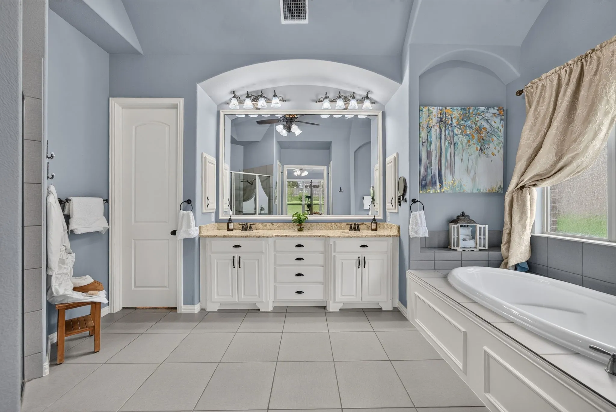Bathroom featuring a dual vanity with white cabinetry and granite countertops, large central mirror, built-in bathtub with tiled surround, and large format tile flooring