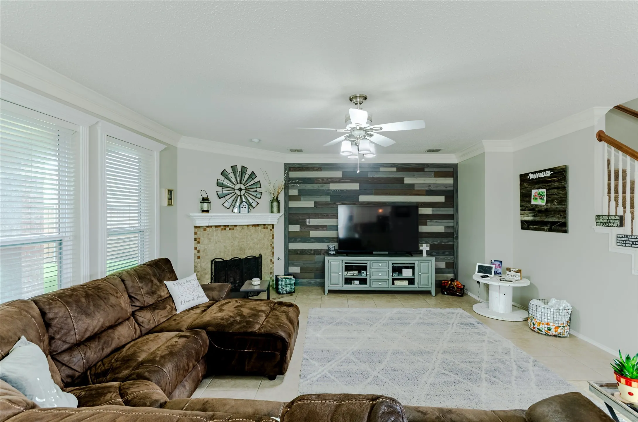 Living space featuring a wood-panel accent wall, a tiled fireplace with hearth, crown molding, multiple windows, and a ceiling fan