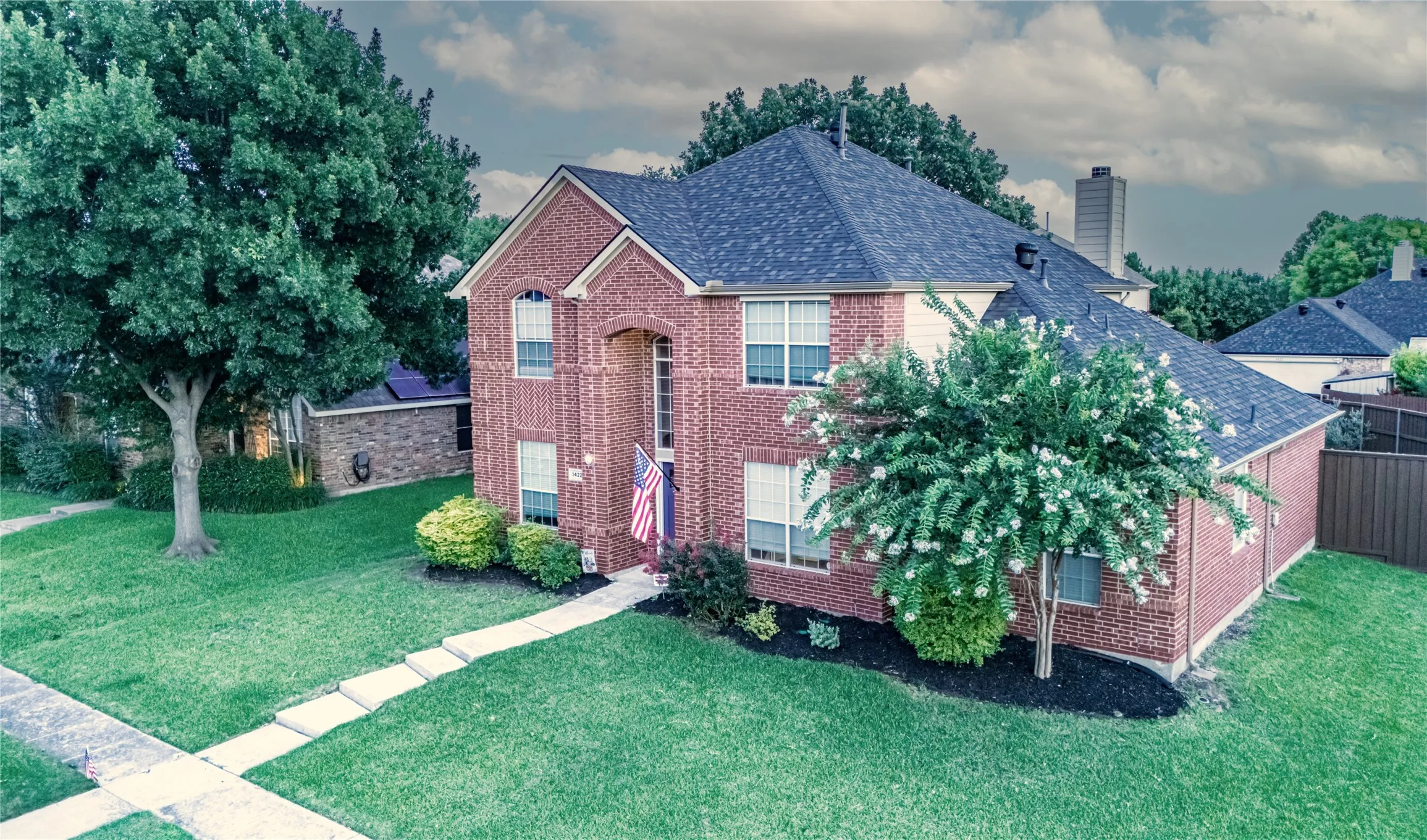 Two-story brick exterior featuring a multi-gabled roofline, mature trees, manicured landscaping, and a side yard with a privacy fence