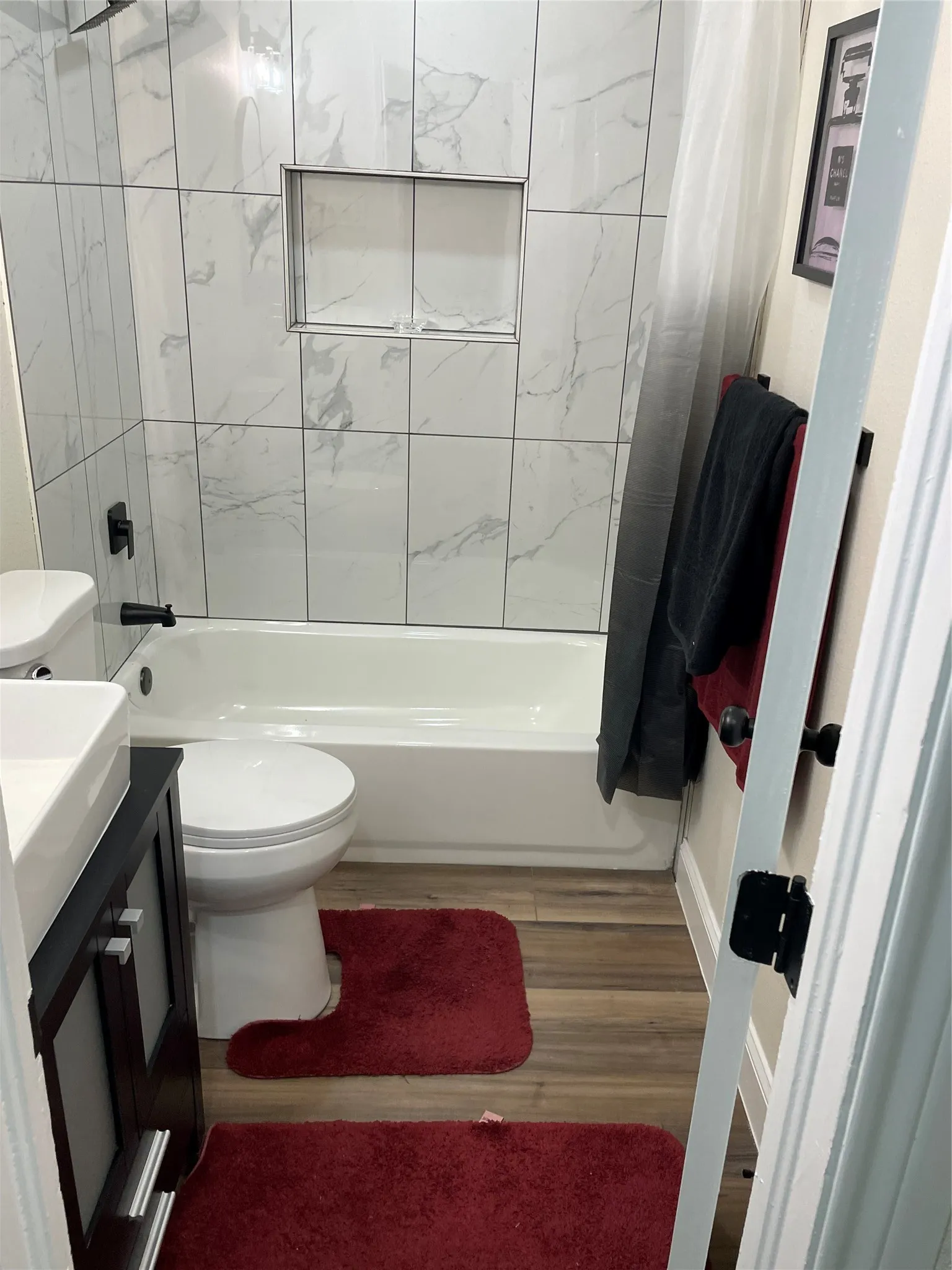 Bathroom featuring a white tub, shower with marble-look tile surround, built-in shower niche, dark wood-finish vanity with square vessel sink, and wood-finish flooring
