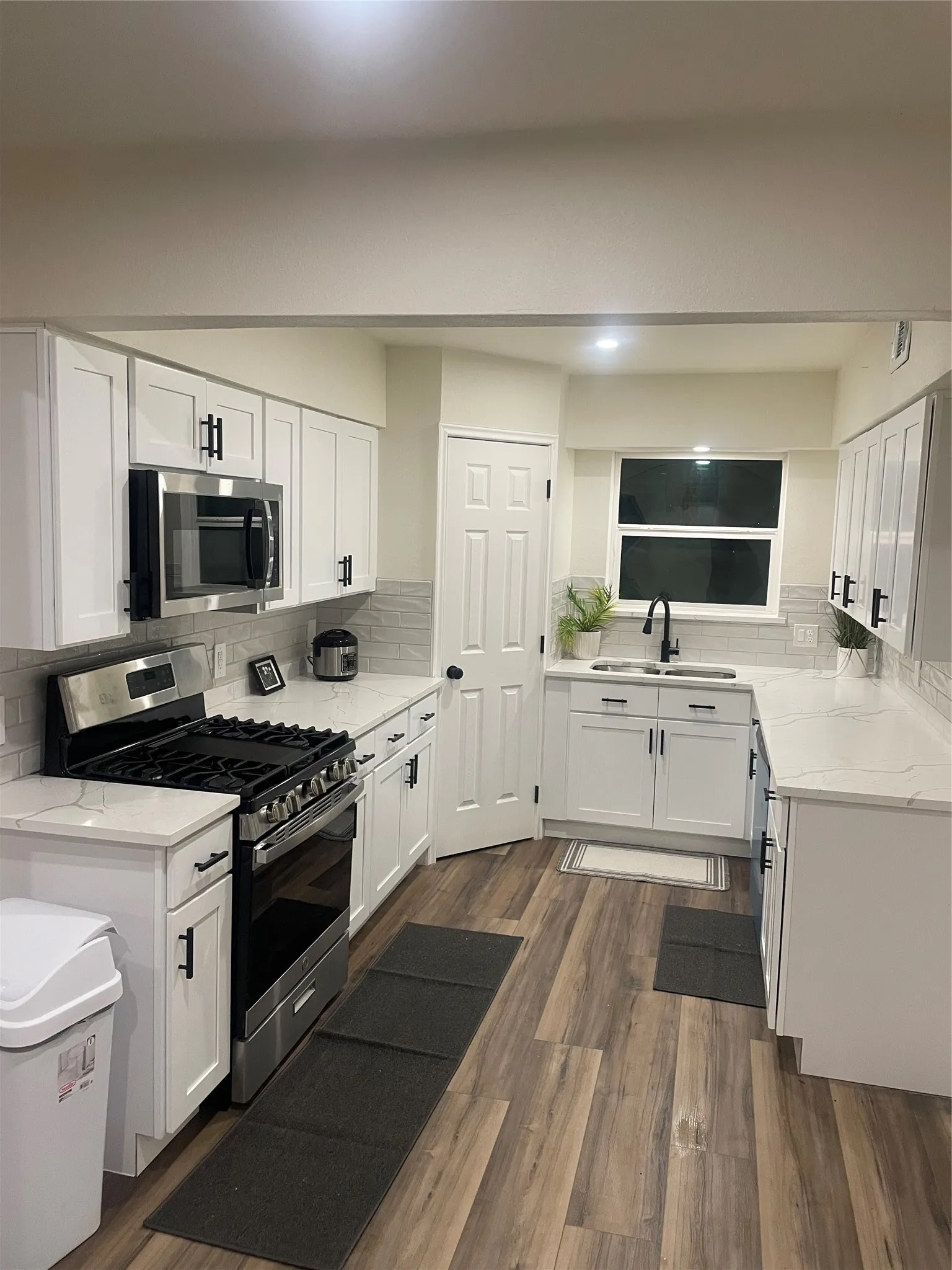 Modern kitchen featuring white shaker cabinetry with matte black hardware, stainless steel appliances, light-toned countertops, a geometric tile backsplash, and wood-finish flooring