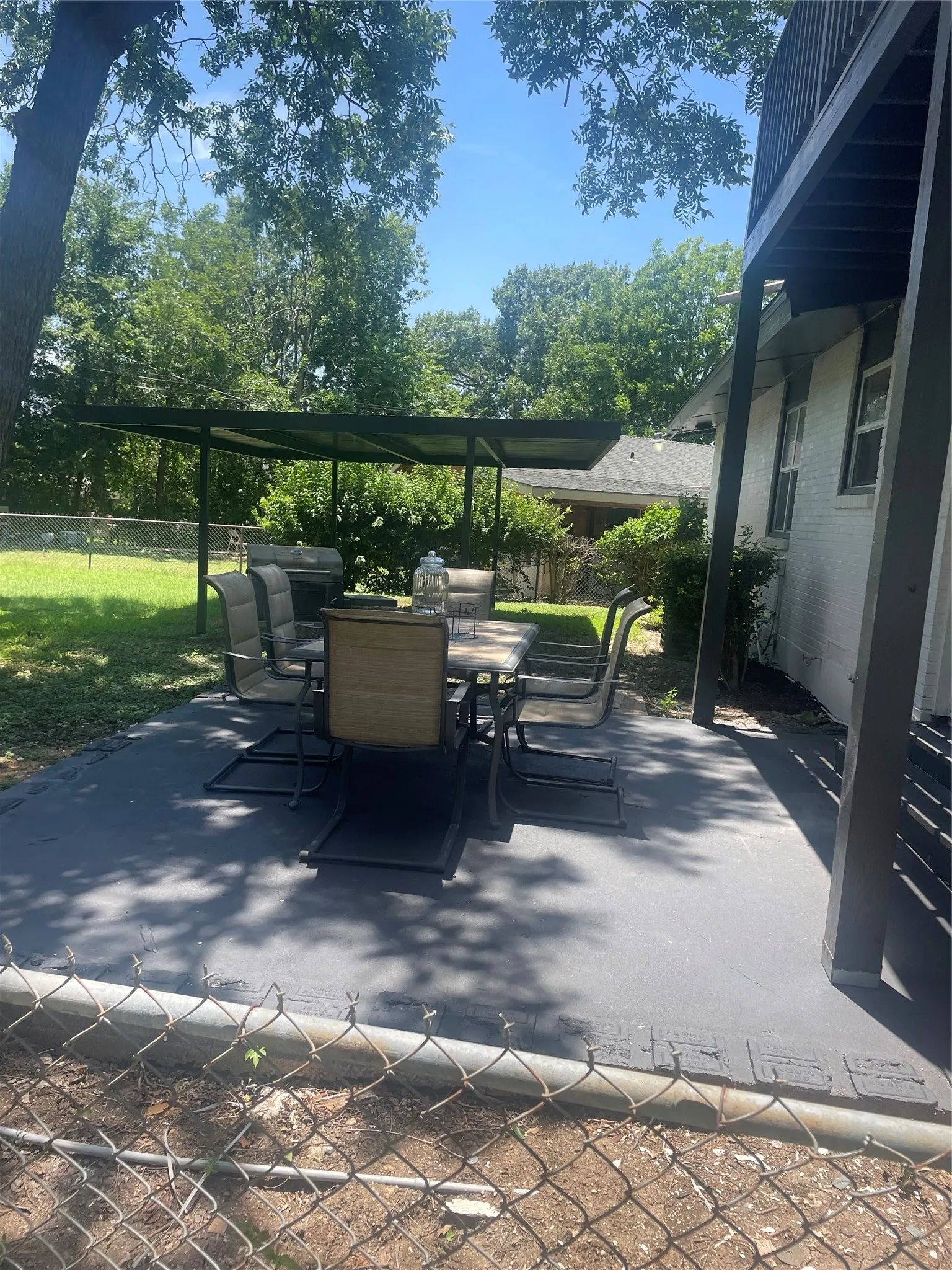 Outdoor patio area featuring a covered dining space, an elevated deck structure, mature trees, and a chain-link fence