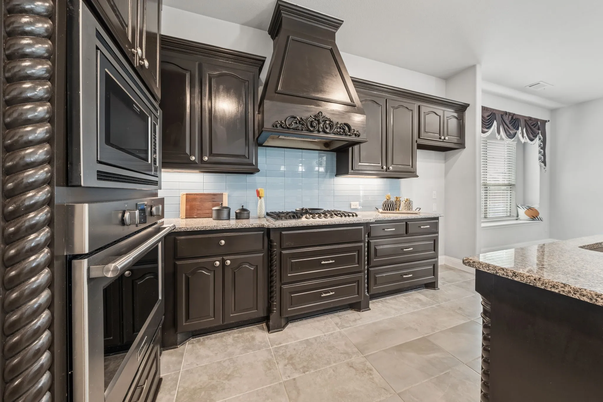 Gourmet kitchen featuring dark wood cabinetry, stainless steel built-in appliances, a decorative range hood, light-colored tile flooring, and a stone countertop island