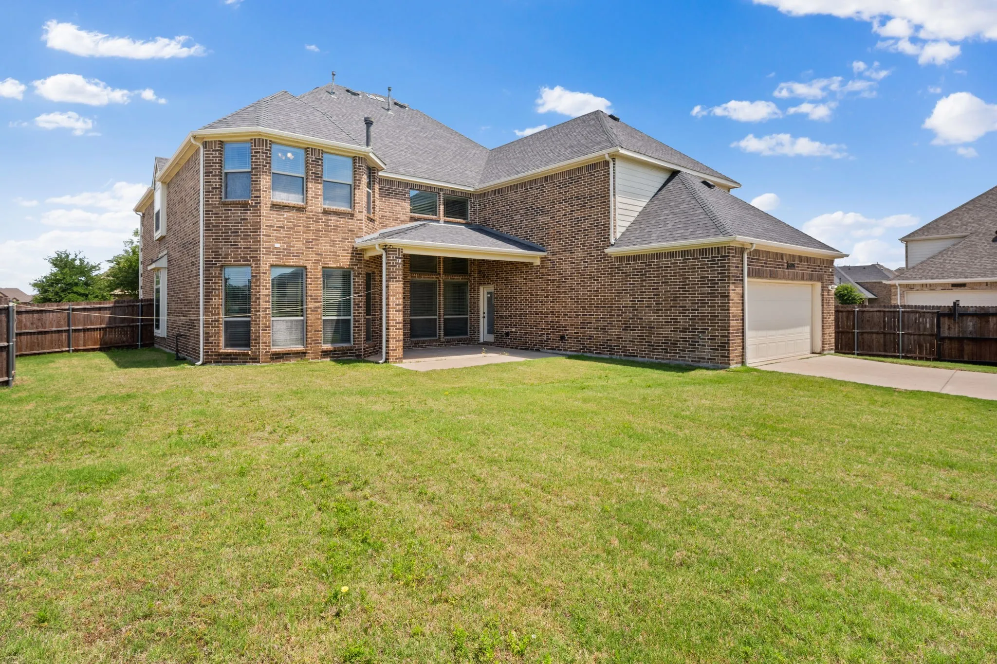Expansive backyard featuring a manicured lawn, full brick exterior, a covered patio, and an attached garage with a concrete driveway