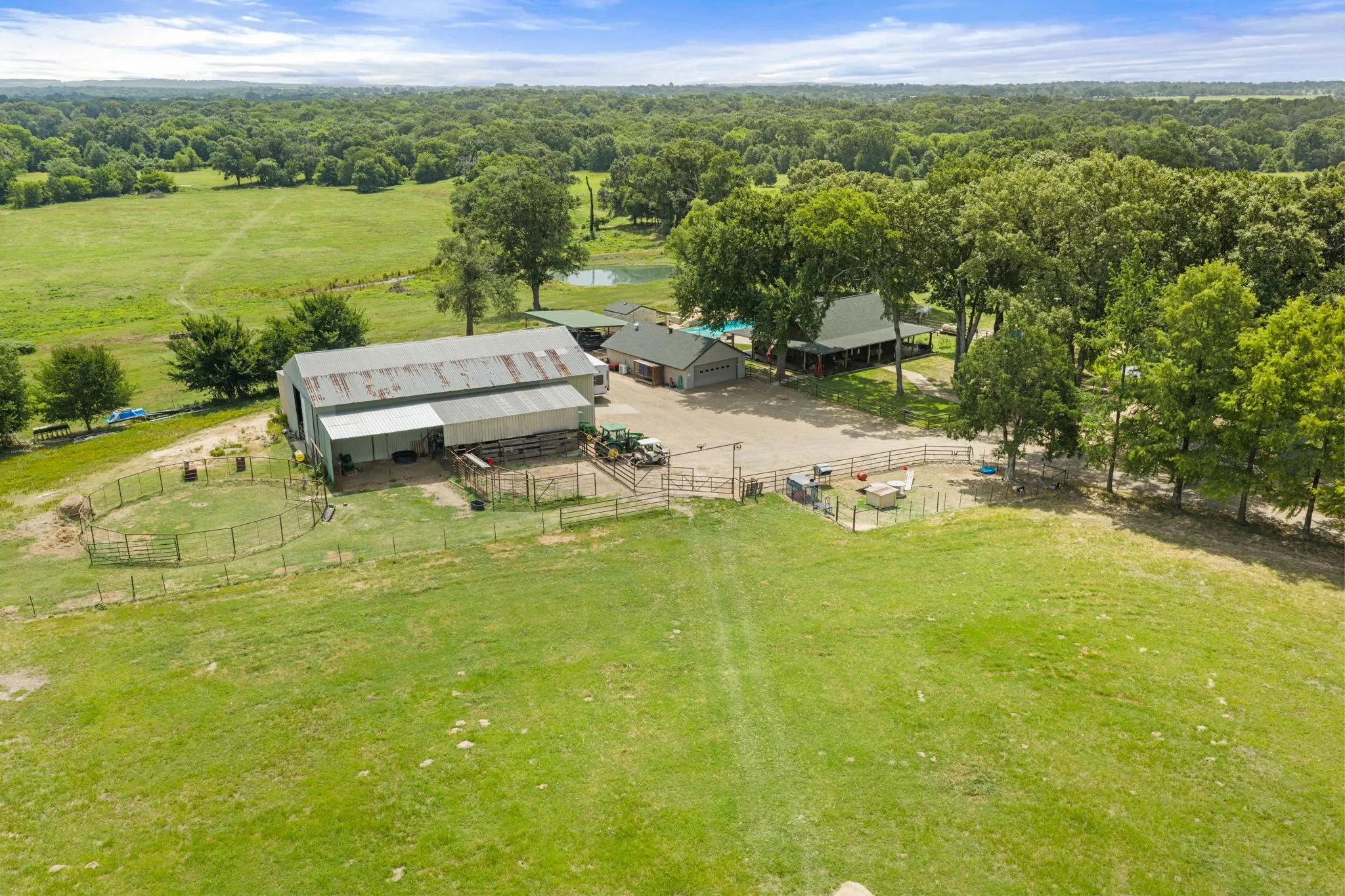 Expansive property featuring a large barn with an attached lean-to, multiple fenced enclosures, a separate building with a green roof, and a visible pond surrounded by mature trees