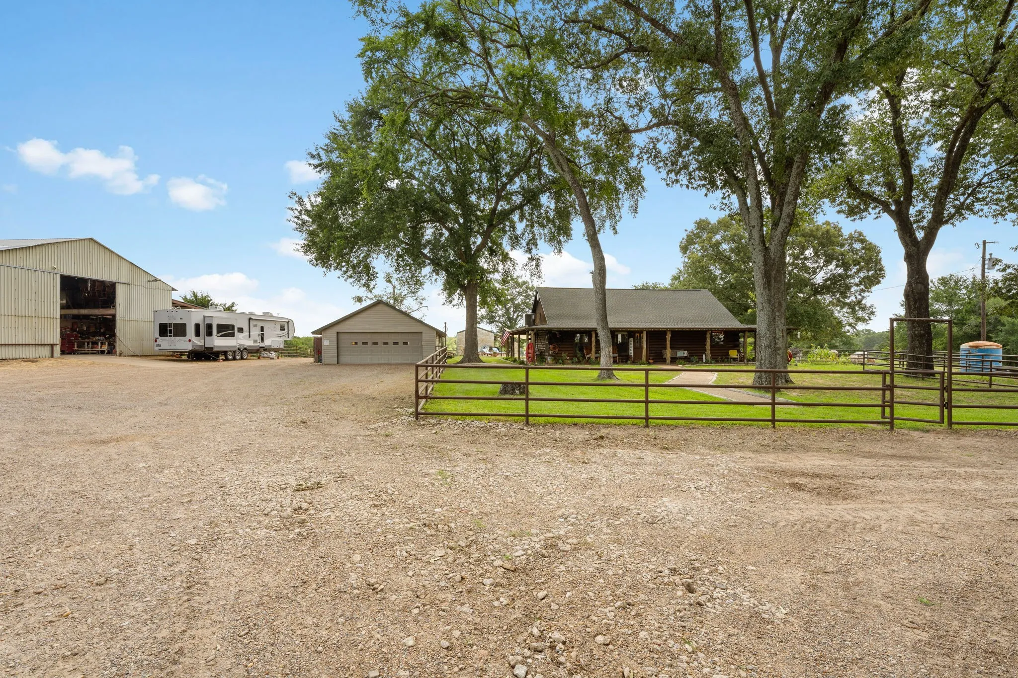 Expansive gravel driveway providing extensive parking, a large metal barn, a detached garage with an overhead door, and a ranch-style home featuring a covered front porch, all surrounded by mature trees and a fenced perimeter