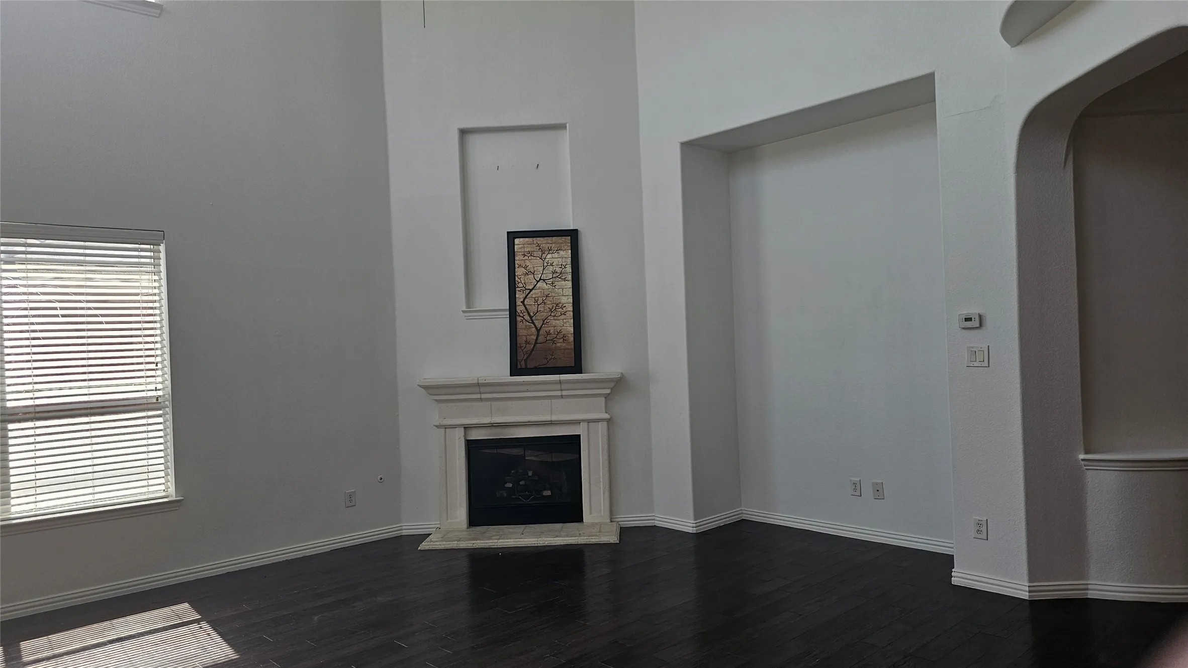 Spacious interior featuring soaring ceilings, a fireplace with a detailed mantel, dark wood-finish flooring, a large window with blinds, and an arched entryway