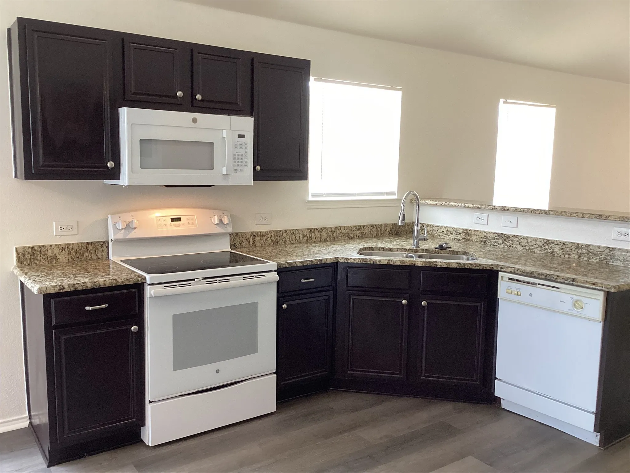Modern kitchen featuring rich dark cabinetry, light speckled countertops, a double-basin stainless steel sink with a high-arc faucet, and white appliances including a built-in microwave, electric range, and dishwasher