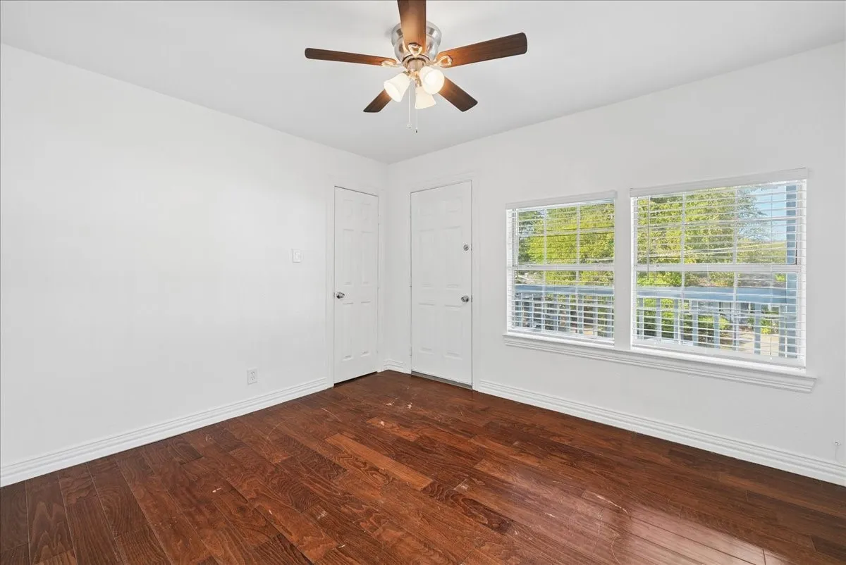 Hardwood-finish flooring, dual windows with blinds, white paneled doors, ceiling fan with light fixture, and white painted walls
