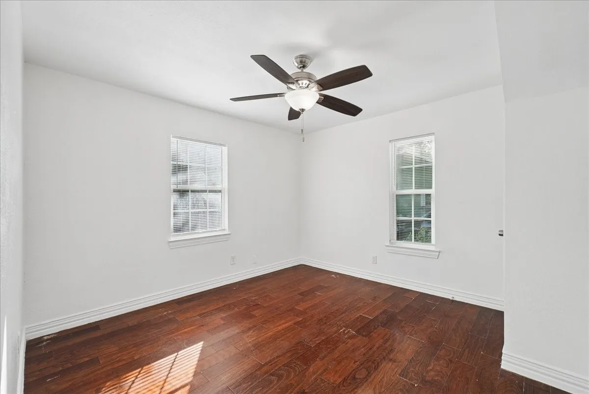 Wood-finish flooring with white baseboards, two windows with white trim, and a ceiling fan with integrated lighting