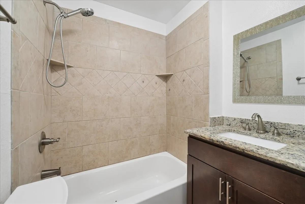 Bathroom featuring a built-in tub with tile surround, a built-in shower shelf, a vanity with a light-toned countertop and integrated sink, a dark wood-finish cabinet, and a framed mirror