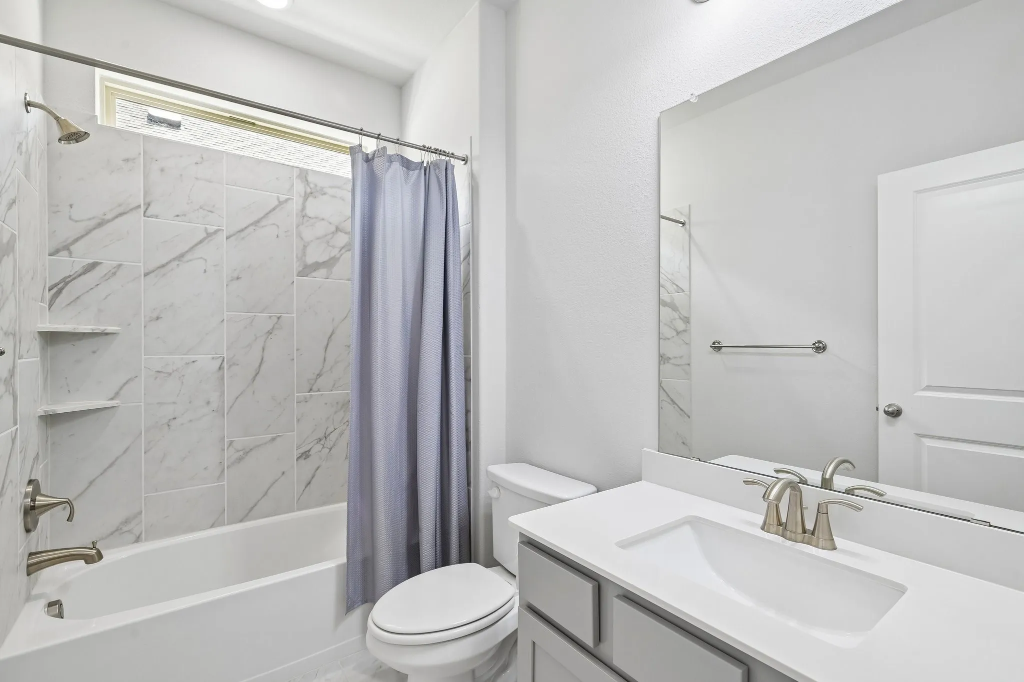 Full bathroom featuring a white tub with white-and-gray marbled tile surround, built-in shelving, and a window for natural illumination